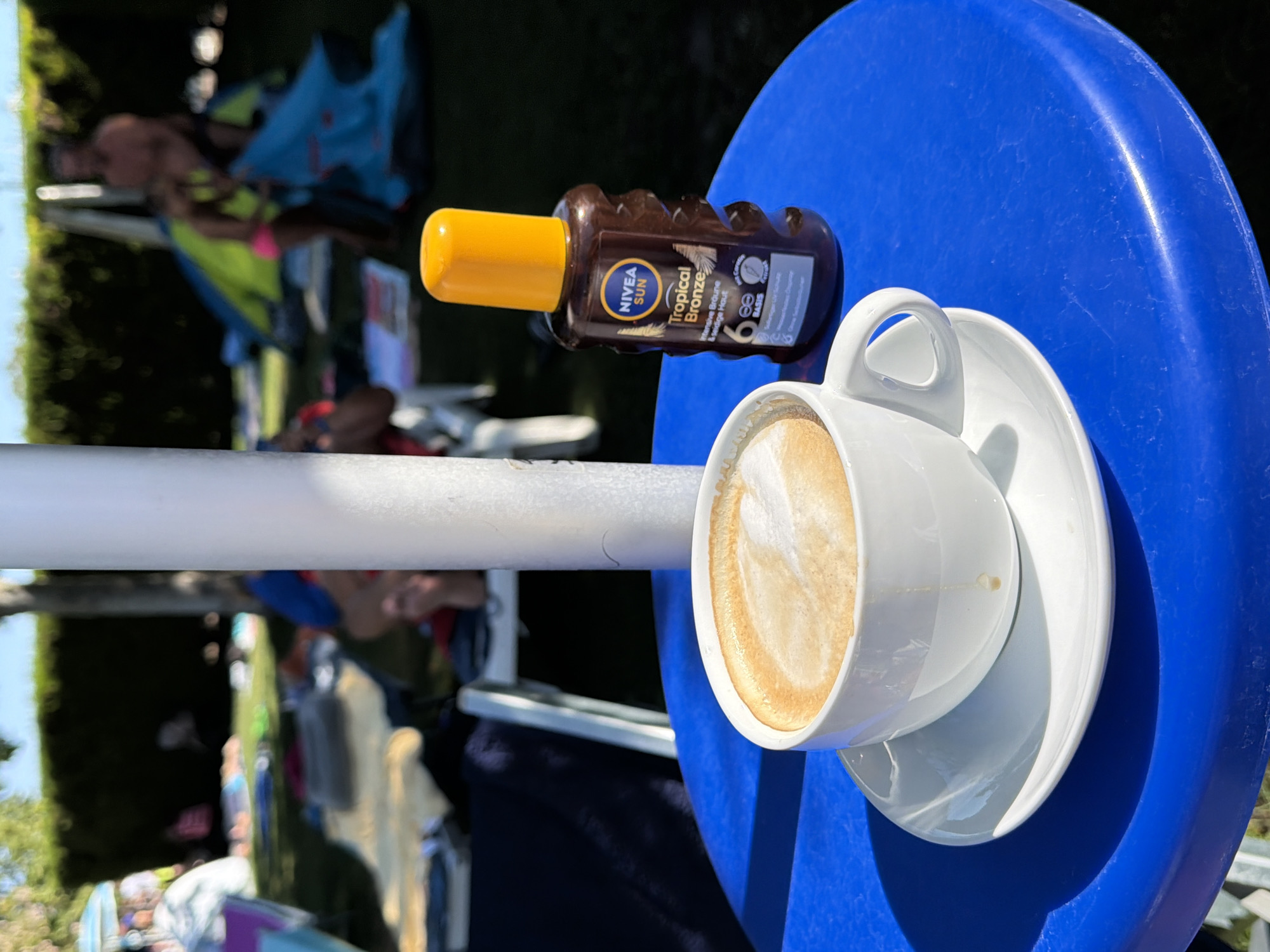 A white cup of coffee with latte art sits on a blue table outdoors, next to a bottle of Nivea Sun Tropical Bronze sunscreen. In the background, people are relaxing on a grassy area with tents and lounge chairs, suggesting a park or camping site setting. The sun is shining, casting shadows. The overall impression is a leisure scene, likely during a warm day.