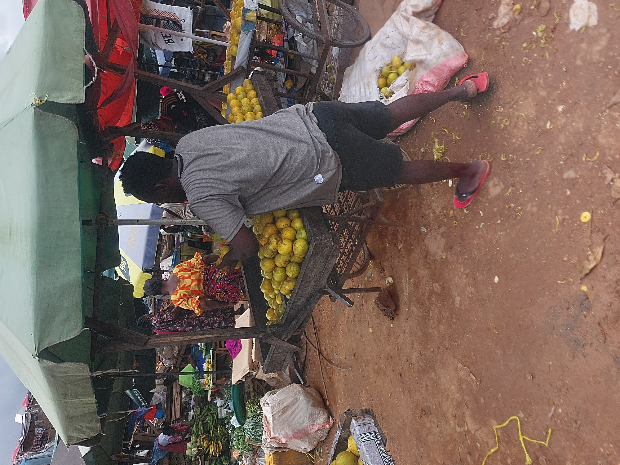 The image depicts a bustling marketplace in Dar es-Salaam, Tanzania. In the foreground, a man wearing a grey t-shirt and black shorts bends over a wooden cart laden with yellow mangoes, seemingly arranging or selecting them. He is wearing red flip-flops.
A woman in a colorful orange and red patterned dress sits nearby, likely a vendor or customer. To the left, a green canopy shades part of the market stall.
The surrounding environment is a dirt surface, with scattered fruit peels and other debris. In the background, there is a variety of fresh produce including green bananas or plantains, along with sacks and other market paraphernalia. Other vendors and customers can be seen in the background, indicating a busy market scene.