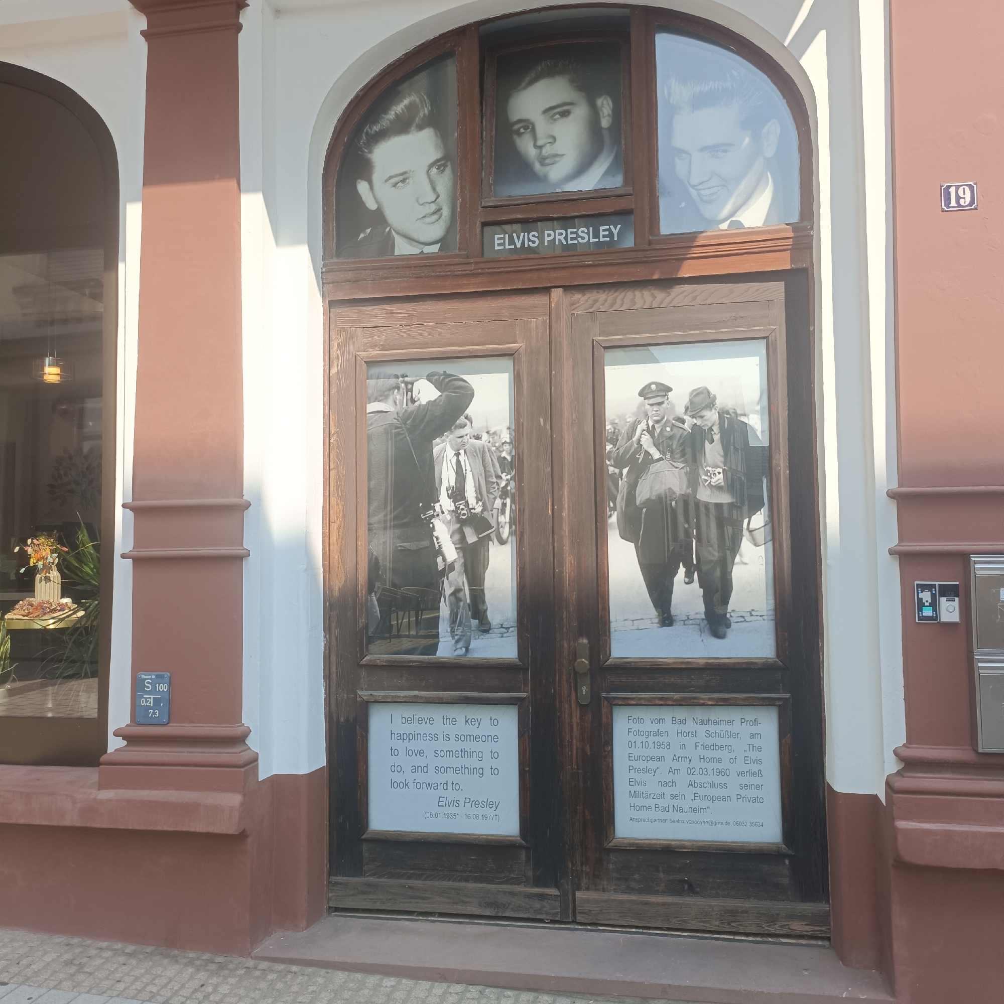 The image shows the entrance to the Elvis Presley Platz in Bad Nauheim, Germany. The entrance consists of a double wooden door with arched glass panels at the top. The arched panels display black and white portraits of Elvis Presley. The word "ELVIS PRESLEY" is visible on a horizontal panel between the arched and rectangular glass sections. Below the arched panels, the doors have two rectangular glass sections each, also featuring black and white photographs. The left door's lower panel displays a quote attributed to Elvis Presley, along with his birth and death dates. The right door's lower panel contains German text detailing a photograph taken in Bad Nauheim on October 1, 1958. The building has reddish-brown and white exterior walls, and a street number "19" is visible on the right wall. The ground in front of the entrance is paved.