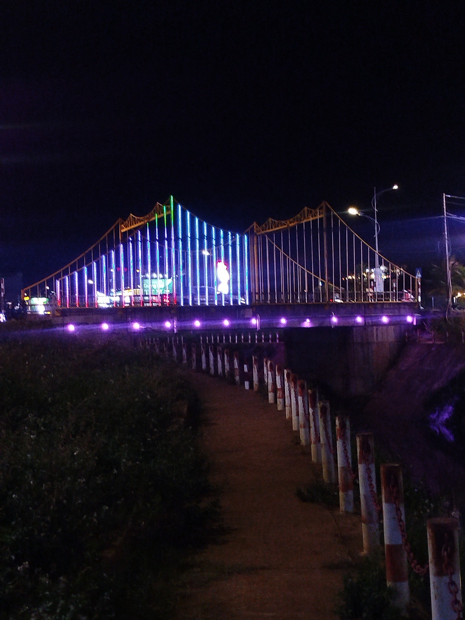 Hoi Phu Ward, Vietnam: A pedestrian path leads to a bridge adorned with blue and green vertical lights. The bridge has an arched design with yellow railings. Purple lights line the base of the bridge. The path is bordered by posts connected by chains. Vegetation is visible to the left of the path, and a street light illuminates the area. The sky is dark.