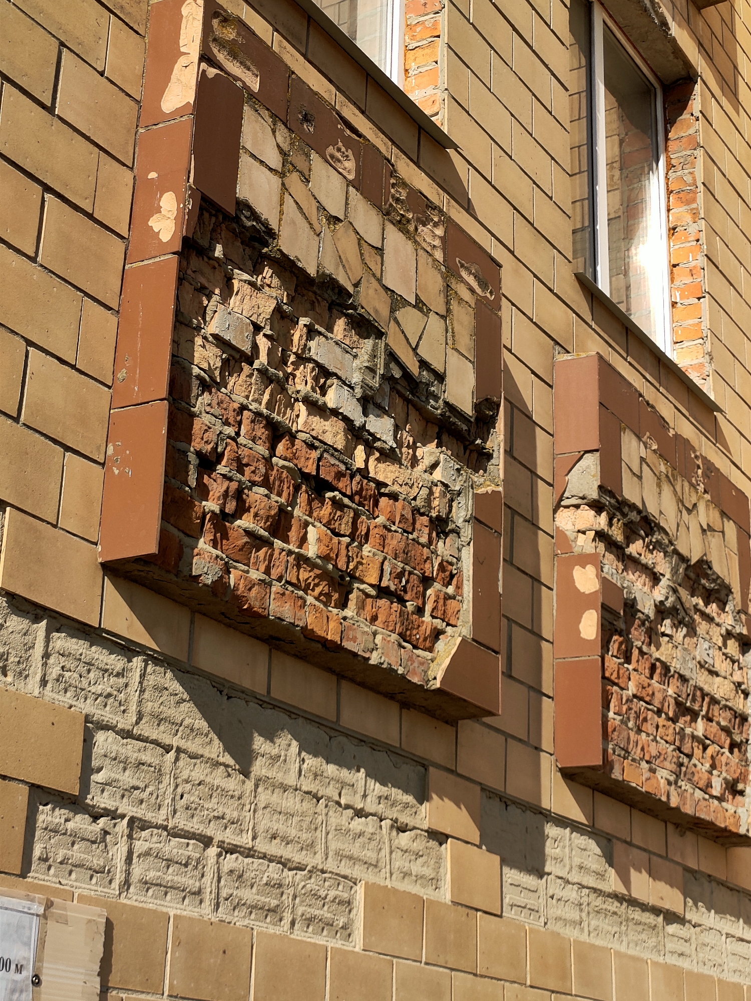 The image depicts a close-up, exterior view of a damaged brick wall of a building, likely in Nosivka, Ukraine. The wall is constructed with light brown brick tiles, some of which are intact and others are broken or missing, revealing the underlying rough brickwork and mortar. Two sections of the wall, one on the left and one on the right, show significant damage. These damaged areas are framed by vertical reddish-brown tile elements. The underlying brick is red and shows signs of age and weathering, with some bricks appearing cracked or crumbled. In the background, part of a window with a white frame is visible, reflecting sunlight. The lower portion of the image shows more intact brickwork, with a lighter, rougher textured stone-like material between rows of brown tiles. The lighting suggests it is daytime, casting shadows on the wall. There are no people or animals visible in the frame.