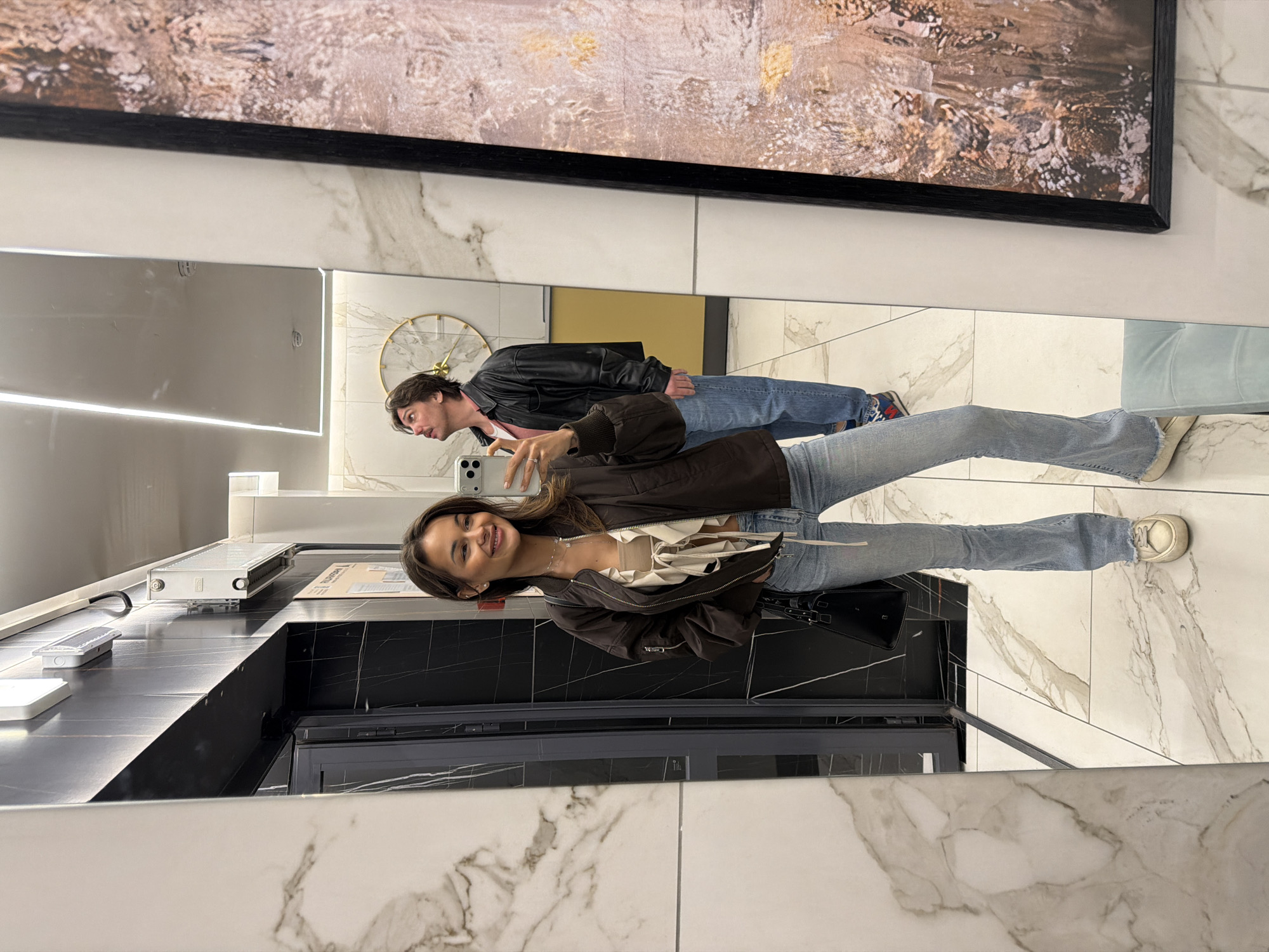 The image captures a mirror selfie featuring two individuals in a modern, well-lit interior. A young woman, presumably the photographer, smiles brightly as she holds a smartphone (identified as an iPhone by its distinct camera array) to capture their reflection. She wears a brown bomber jacket unzipped over a light-colored, possibly white, top with tie details, and blue flared jeans with white sneakers. A black handbag rests on the floor beside her feet.
Behind and to her left, a young man stands, also reflected in the mirror, looking towards the camera with a neutral expression. He is dressed in a black leather jacket over a reddish or pink shirt and blue jeans.
The setting appears to be a sophisticated indoor space, characterized by a large rectangular mirror. The walls and floor are covered in elegant white marble with subtle grey veining, contrasting with sections of dark, possibly black, tiles or countertop surfaces. A long, dark countertop runs in the foreground, equipped with linear LED lighting strips. On the countertop, various items are visible, including a white power strip or electronic device, another white rectangular object, and a document displaying partially visible Russian text, "РУС".
Above the main mirror, a large, rectangular framed abstract artwork with earthy tones and textures hangs on the wall. Further back on the wall, behind the man, a circular gold-framed wall clock with Roman numerals adds a touch of classic design. The overall impression is a casual moment captured in a stylish environment, possibly in Krasnoyarsk, Russia.