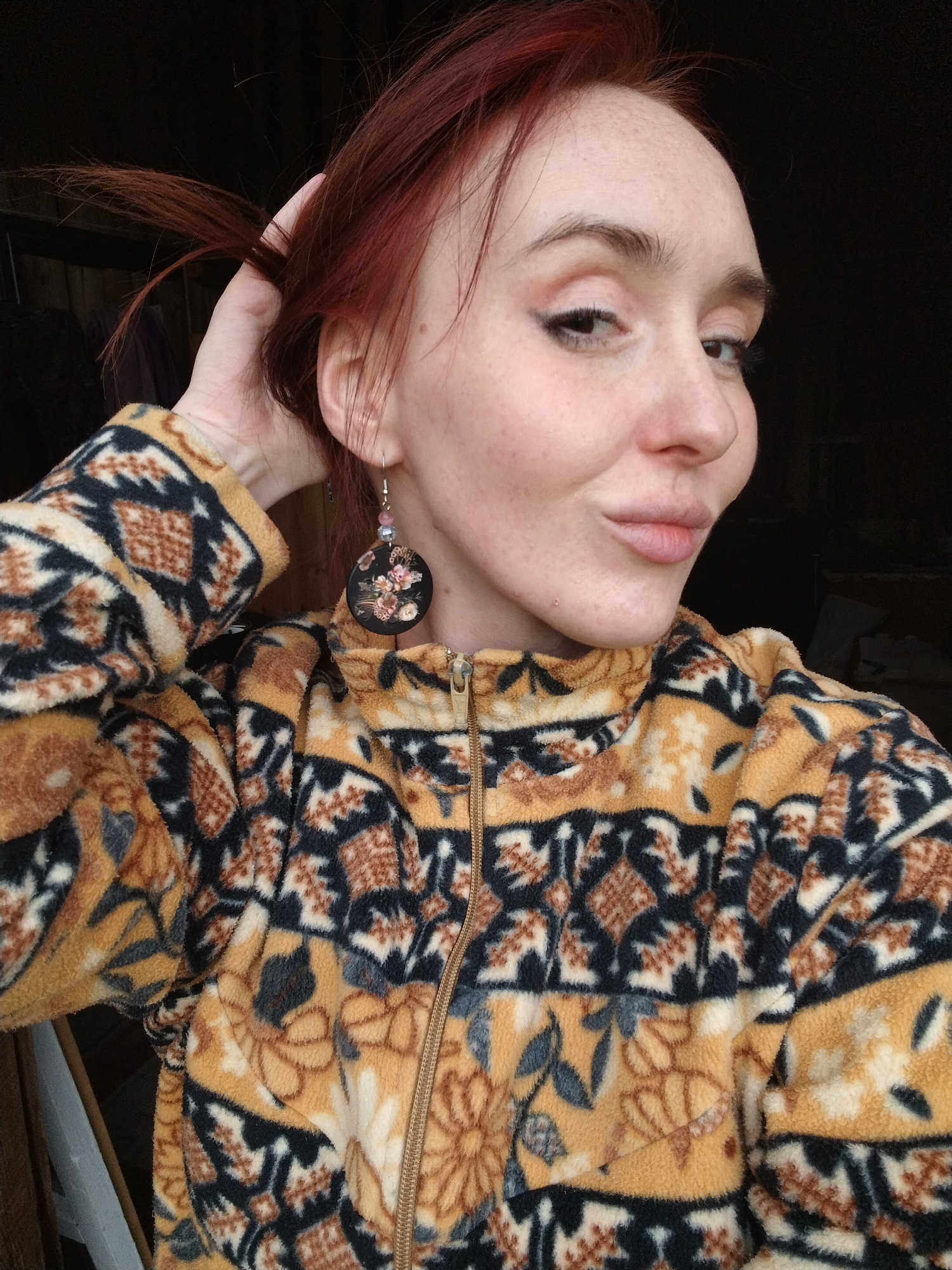 A woman with fair skin, red hair, and freckles poses for a close-up selfie. She is wearing a fleece jacket with a yellow and brown geometric and floral print. Her left hand is raised, holding a few strands of her hair near her ear. She is wearing a dangling earring on her left ear featuring a black disc with a floral design. The background is dark and out of focus, but appears to be an interior space. There is no verifiable information about the city of Krasnogorskoye, Russia in the image.