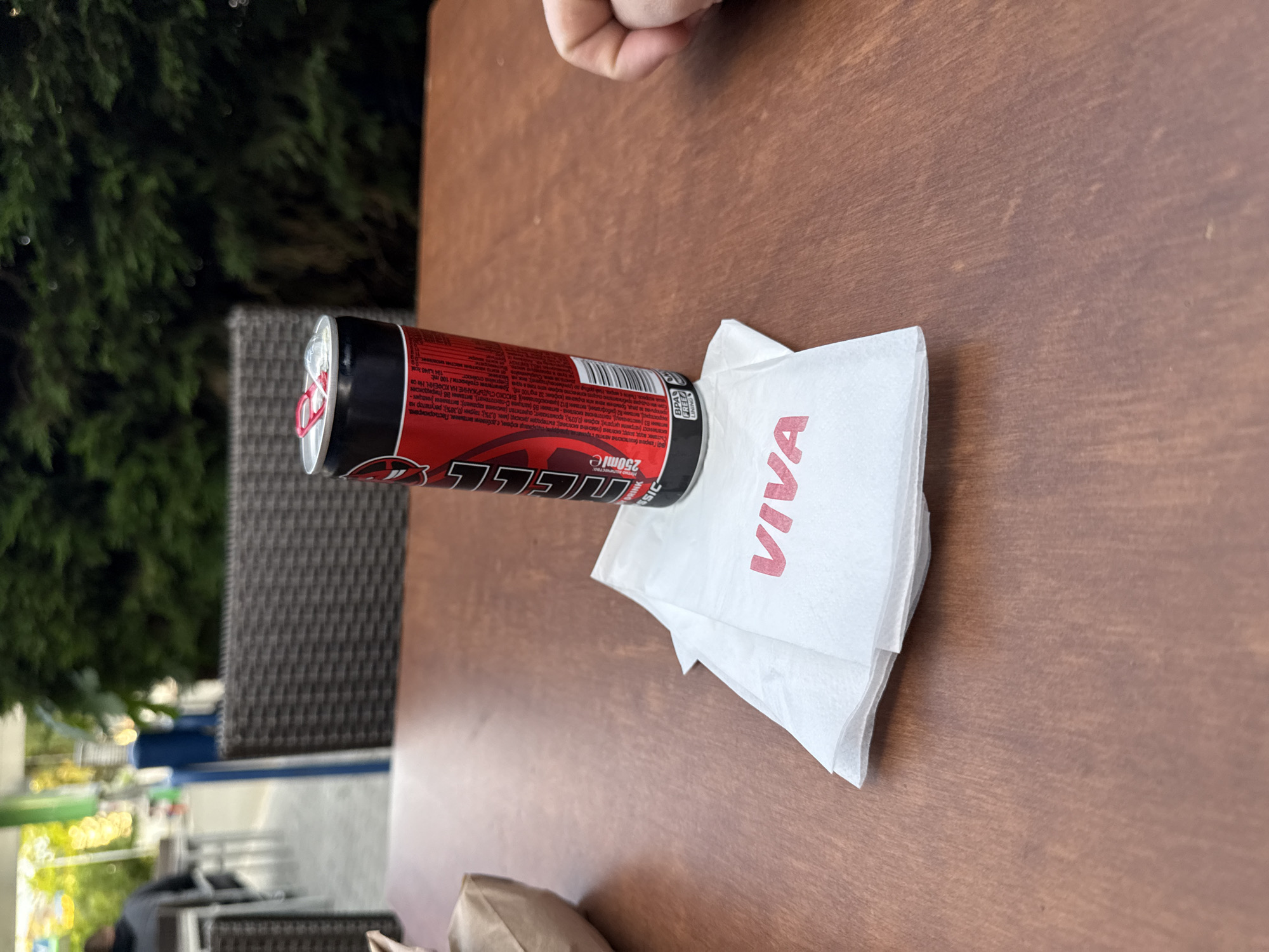 A can of "Energy Classic" VIVA energy drink rests horizontally on top of several white paper napkins with the word "VIVA" printed in red letters on the table. The can is red with black accents, and a silver top. The table is brown and wooden, with a person's hand visible in the upper right corner of the image. In the background is a woven chair and greenery that seems to be outside. The image appears to be taken at a cafe or outdoor seating area. Based on the VIVA branding and information in the OCR, this image appears to be from Stara Zagora, Bulgaria.
