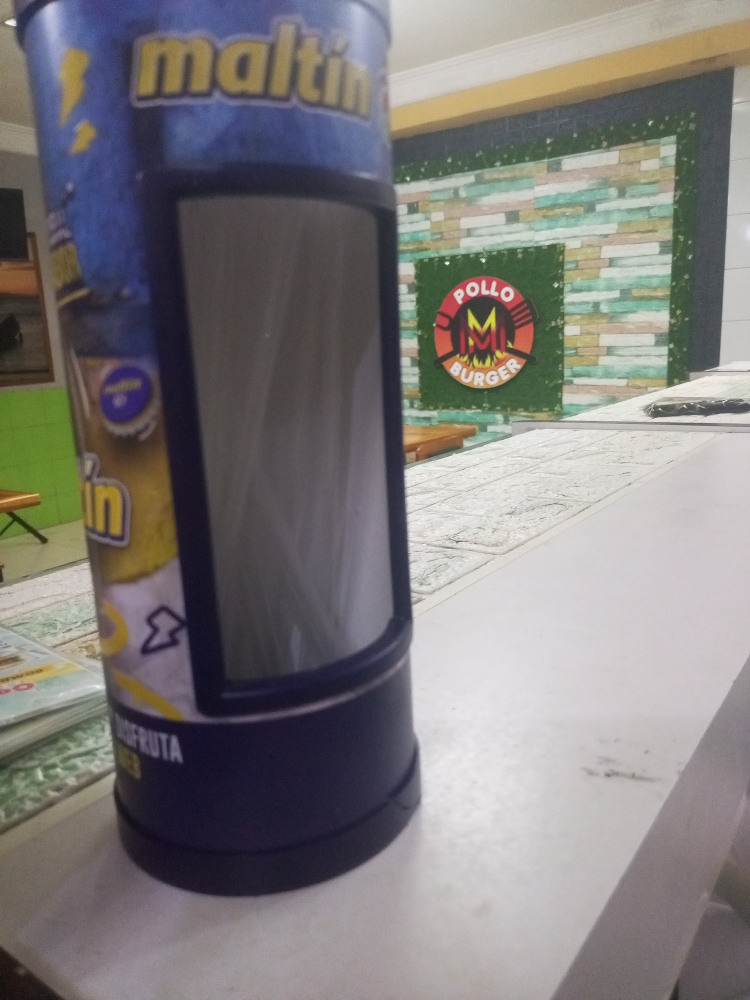 The image shows a Maltin Power can on a white counter. In the background, a "Pollo M Burger" sign is visible on a wall with a decorative brick pattern and green foliage. Based on the sign and product, the location is likely a restaurant in Juan Griego, Venezuela.