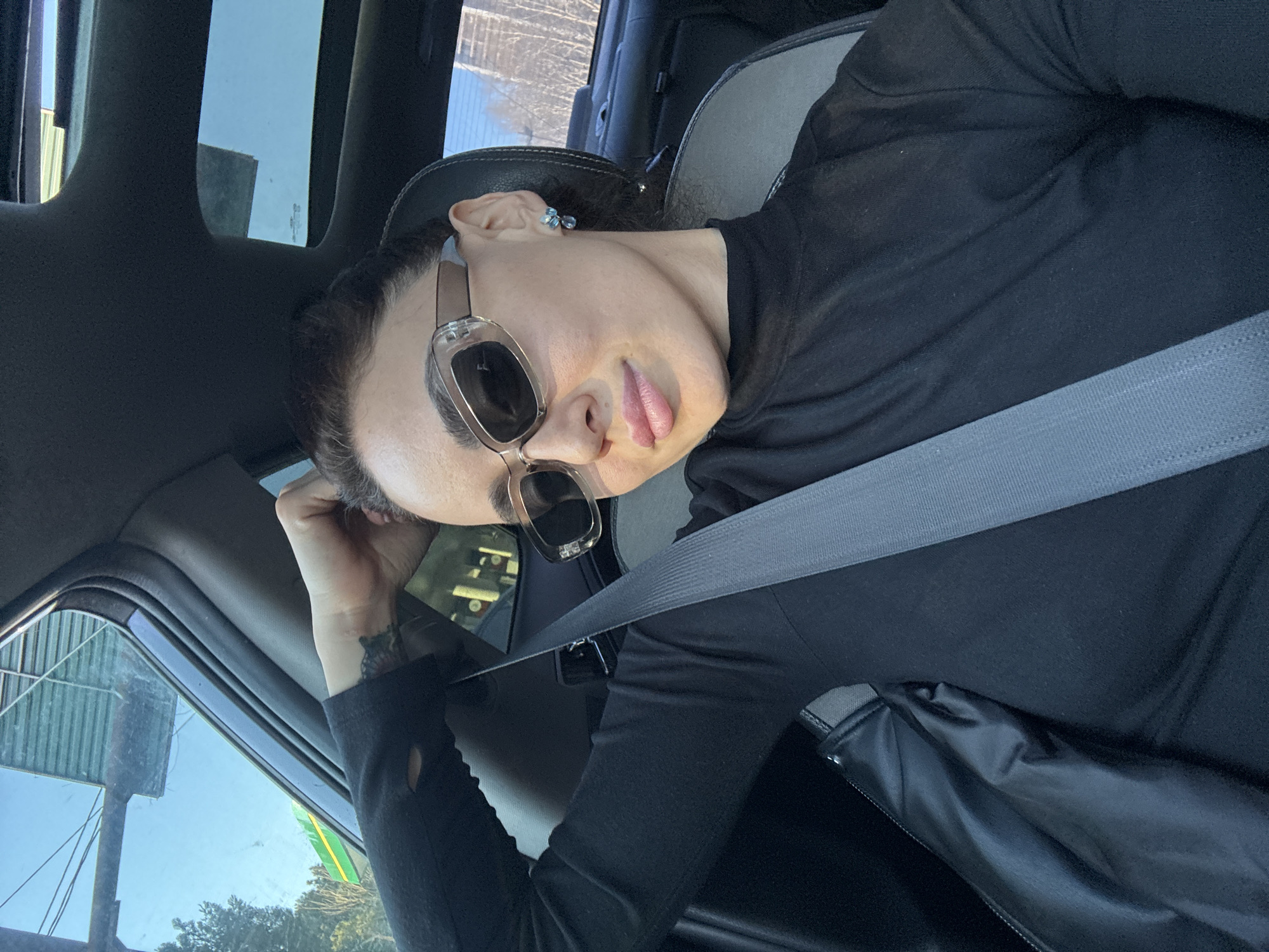The image captures a female individual seated in the front passenger seat of a vehicle during daytime in Tbilisi, Georgia. She wears a black long-sleeved turtleneck, rectangular framed sunglasses, and light blue drop earrings. Her dark hair is styled back, and her right hand is positioned near the back of her head. A gray seatbelt secures her across the torso. A small, dark tattoo is visible on her inner right wrist. The vehicle's interior features dark upholstery, a visible sunroof, and a black and gray accented seat.
The exterior environment, visible through the vehicle's windows, places the scene in an urban area of Tbilisi, Georgia. Through the driver's side window, a clear blue sky, bare deciduous trees, and a large green and yellow corrugated structure, identifiable as a Wissol gas station canopy, are evident. Through the rear window, further bare trees and modern high-rise buildings are discernible against a clear blue sky. The scene is illuminated by strong natural sunlight.