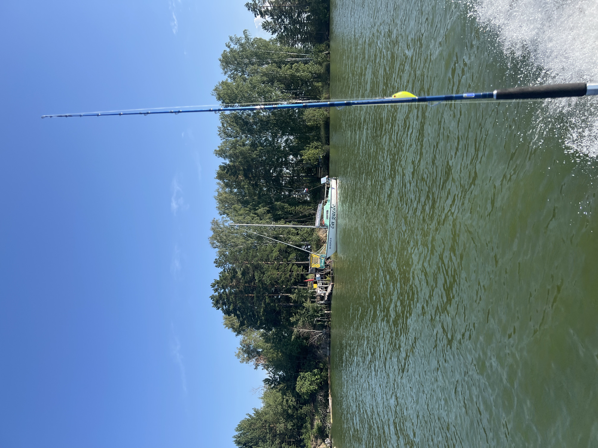 The image depicts a serene daytime scene on a body of water, likely a lake or river, with a shoreline lined with lush green trees. In the foreground, a fishing rod is prominently displayed, extending upwards from the bottom right corner of the frame. A yellow bobber is attached to the fishing line. The water is a murky green color, with small waves indicating movement, possibly from a boat. In the middle ground, a white sailboat with a green canopy is anchored near a wooden pier. The pier appears to be part of a small settlement or recreational area along the shore. The background consists of a clear blue sky with a few wispy clouds. The location is identified as Borovoe, Russia, suggesting a natural setting often associated with outdoor activities like fishing and boating. There are no people visible in the image. The overall atmosphere is tranquil and suggestive of a peaceful day spent outdoors.