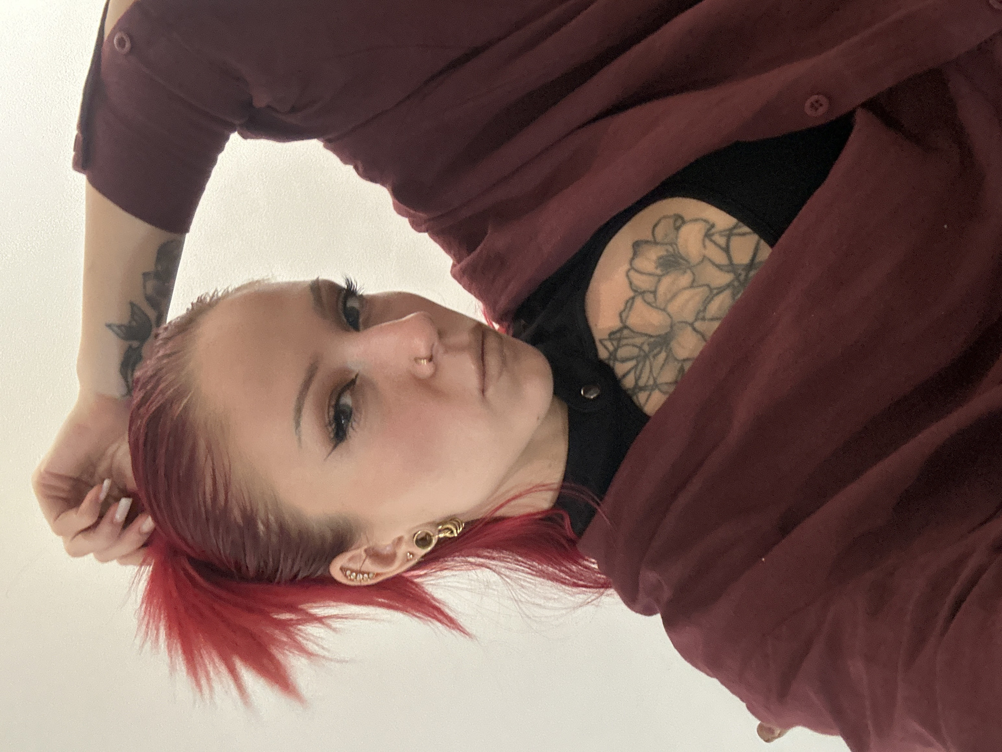 A woman with red hair is shown in a close-up shot. She is wearing a maroon button-down shirt over a black top. She has tattoos on her chest and left arm. She also has multiple ear piercings. The background is a plain white wall. There is no indication of the image being taken in Kerobokan Kelod, Indonesia.