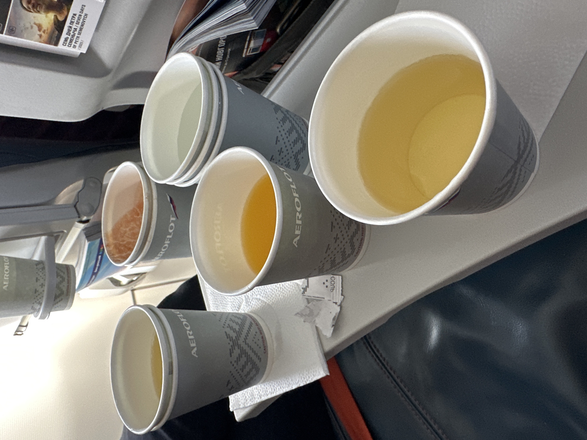 The image depicts a tray table on an airplane, likely in economy class, with four cups of beverages. Three of the cups are partially filled with liquid, one with a bright orange juice, and two with lighter yellow liquids, possibly tea or water. The fourth cup is empty and stacked on top of another empty cup, suggesting these are for use. The cups are grey with a geometric pattern and the "Aeroflot" logo. There is also a white paper napkin and a small packet of sugar. In the background, a magazine is visible with a picture of a man's face and Russian text. The seats in front and beside are visible, one of which is upholstered in dark material. The scene is well-lit, likely by interior cabin lighting, and the overall impression is one of a standard in-flight service. There is no specific indication of the location being the city of Sadovoe, Russia, but given Aeroflot is the national airline of Russia, it is plausible.