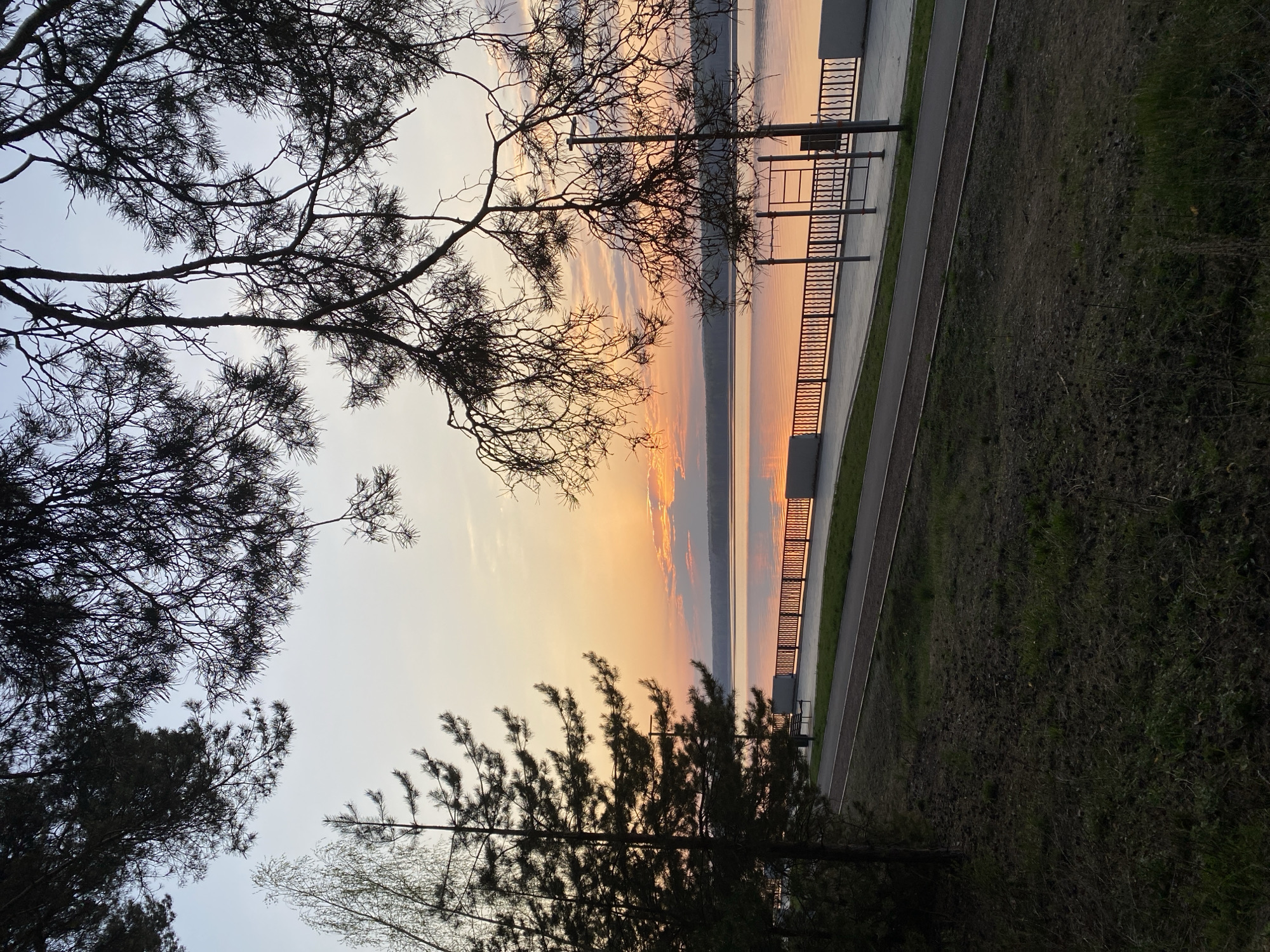 The image depicts a sunset view in Chaikovsky, Russia, overlooking a body of water. In the foreground, silhouetted trees frame the scene. Adjacent to the water is a paved walkway with a metal railing. Beyond the walkway is a grassy area. The sky exhibits colors from orange to light blue. There are no people visible.