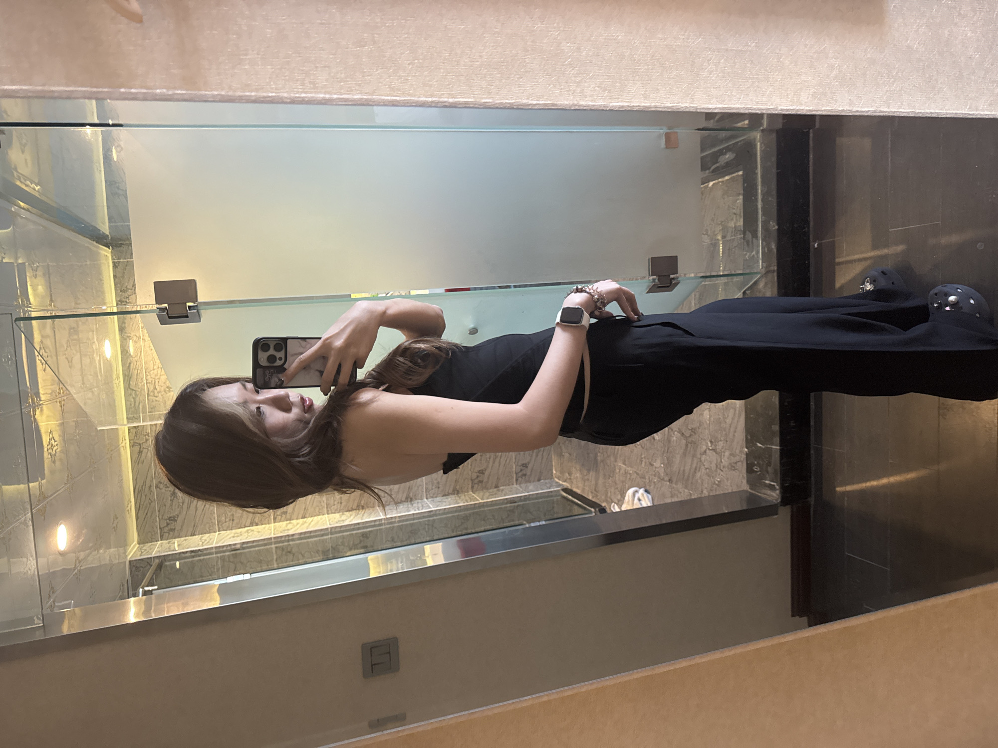 A young woman is depicted in a mirror selfie, taken indoors within what appears to be a modern bathroom or hotel room in Ganzhou, China. She is positioned upright, holding a dark-colored smartphone with a light-colored, possibly marble-patterned case, in her right hand, angled to capture her reflection. Her gaze is directed towards the phone screen, and she has a neutral expression with slightly parted lips.
She has long, straight, dark brown hair with lighter highlights. Her attire consists of a black sleeveless top and black trousers. On her left wrist, she wears a white smartwatch and multiple silver-toned bracelets. Her feet are visible at the bottom of the frame, wearing black slip-on shoes, possibly Crocs, adorned with small, sparkling embellishments.
The reflected background reveals a contemporary interior design. A wall with light-colored, patterned tiles is visible behind her, featuring a small circular light fixture. A clear glass partition, possibly a shower enclosure or room divider, with metal hinges, traverses the middle of the scene. Below this glass, there is a dark-colored counter or ledge, and further back, darker, possibly marble-patterned, wall or floor tiles are discernible. Faint reflections of white objects, possibly towels, can also be seen through the glass. The upper portion of the reflection shows a textured, light-colored surface, likely a ceiling or a large lampshade. The lighting is artificial, characteristic of an indoor setting, and there is no visible text in the image.