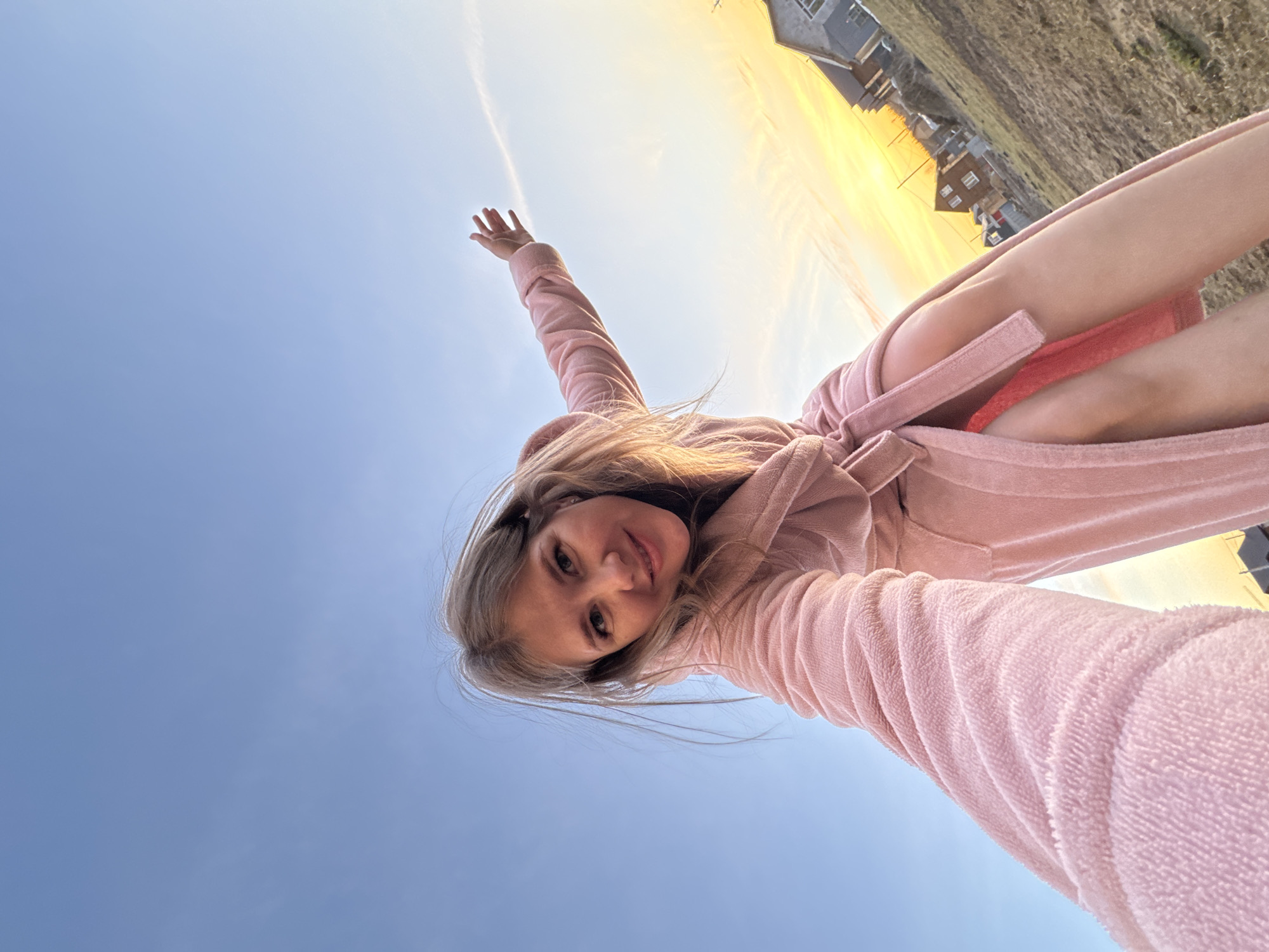 The image is a low-angle shot of a young woman wearing a pink robe. She is looking down at the camera with her left arm raised towards the blue sky. In the background, a residential area with houses is visible under a golden sky. The location is Gornyi, in the Dvurechenskoe S/P area of Russia. The sky is a clear, bright blue. There's a light cloud in the background. The woman's robe is tied at the waist.