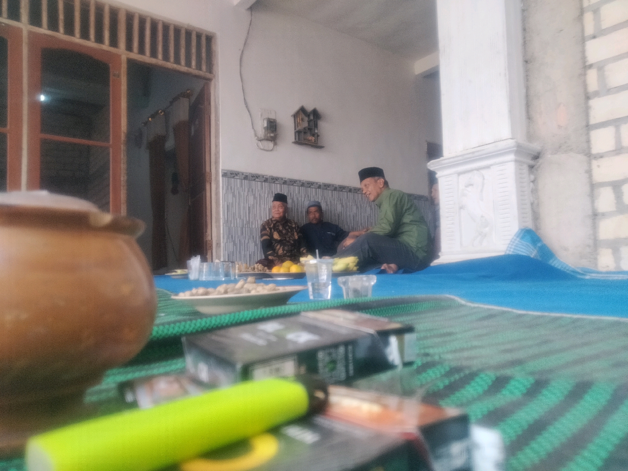 The image depicts an outdoor gathering at a residence in Soko, Indonesia. In the foreground, a large, round, brown ceramic pot is prominently displayed. In front of it, a bright green cylindrical object rests on a mat with a green and black striped pattern. Several packs of cigarettes are scattered on the mat as well.
In the mid-ground, three men are seated on a blue mat. The man on the left wears a black peci hat and a patterned shirt. The man in the center also wears a peci hat and a dark shirt. The man on the right, wearing a green shirt and a peci hat, is engaged in conversation with the other two. A plate of food and drinks are visible on a smaller white plate to their left.
The background reveals the exterior of a building with wooden doors and window frames. A decorative shelf with a small house-shaped object and an electrical meter are mounted on the wall. The overall atmosphere appears to be a casual social gathering.