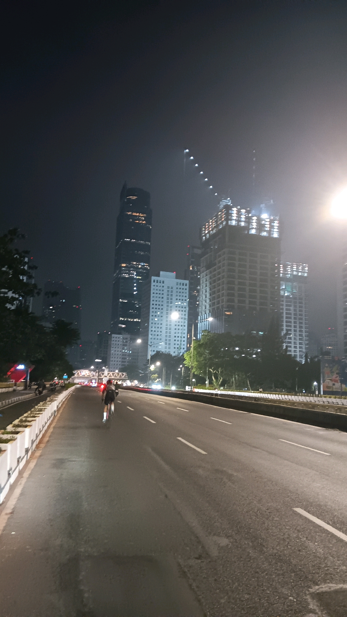 The image shows a nighttime scene on a multi-lane road in a city. In the foreground, a person is cycling on the right side of the road. The road surface is wet and reflects the light from street lamps and vehicle headlights, which are blurred and create streaks of light. Several cars and motorcycles are visible in the distance, their lights creating red and white streaks. The left side of the road is bordered by a concrete barrier with white rectangular planters.
In the background, a dense collection of high-rise buildings dominates the skyline. Some of these buildings are illuminated, showing a pattern of lit windows. Several buildings appear to be under construction, with scaffolding and cranes visible. The sky is dark and hazy, with some lights from the city piercing through the atmospheric haze. The overall impression is one of a bustling urban environment at night, possibly during a period of development or renovation. The presence of the cycling person and vehicles suggests the road is open for traffic and recreational activities. The scene is likely captured in an area with significant urban development, characteristic of a major city. Considering the architectural style of the buildings and the overall urban density, this scene could be located in a business district or a rapidly developing area of a major metropolitan city.