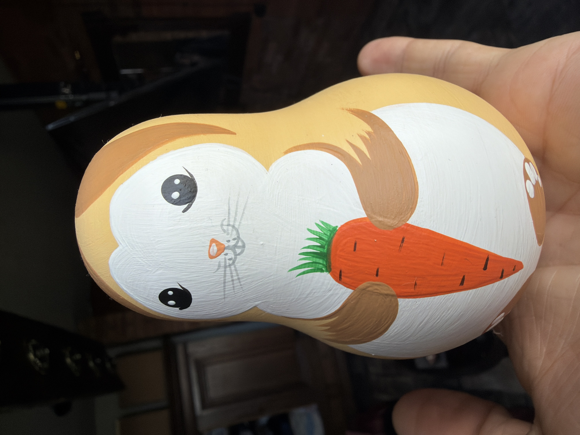 A hand is holding a brightly painted wooden nesting doll shaped like a rabbit holding a carrot. The doll is primarily white and orange, with brown accents for the rabbit's paws and ear outlines. The carrot is orange with green leaves on top. The background is dark and out of focus, showing hints of indoor furniture and lighting fixtures. The doll is positioned in the center of the image, filling most of the frame.