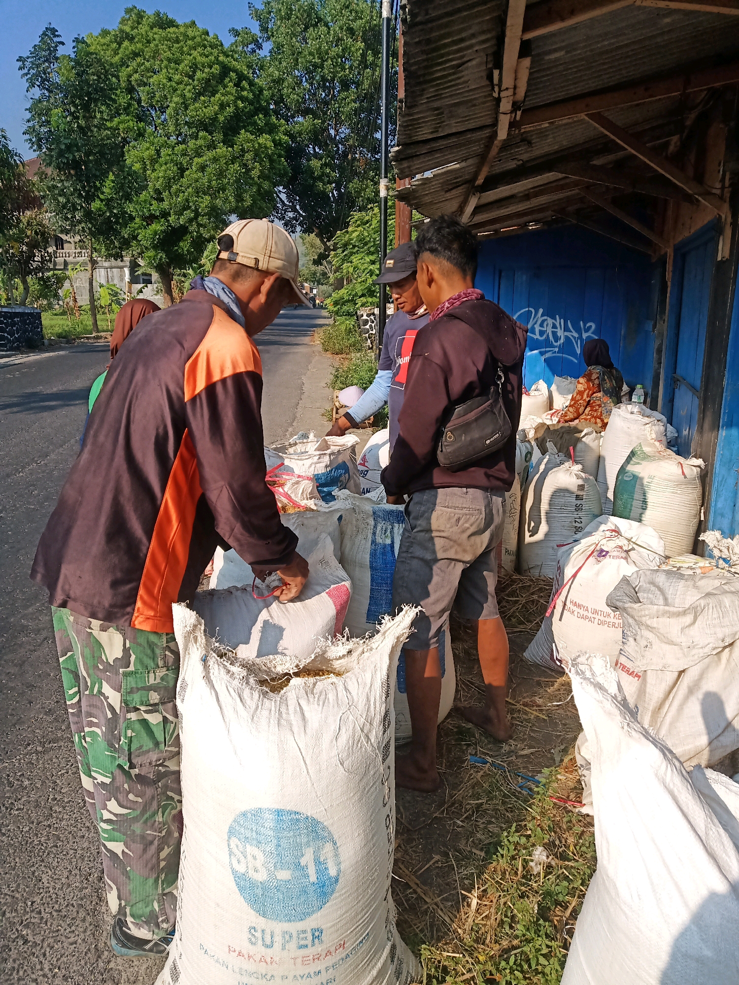 The image depicts an outdoor scene likely in Garut, Indonesia, with three individuals engaged in activity around large sacks of goods. Two men are standing in the foreground, one wearing a brown and orange long-sleeved shirt and camouflage pants, and another in a dark hooded sweatshirt and grey shorts. The man in camouflage is holding a pair of red-handled scissors and appears to be interacting with the opening of a large white sack in front of him. The man in the sweatshirt has a small black bag slung across his chest and is looking towards the sacks. A third individual, wearing a dark cap and a blue shirt, is partially visible between them, also near the sacks. Behind the men, several more large sacks are piled up, some with visible writing. The sacks are positioned next to a structure with a corrugated metal roof and blue wooden walls. The environment appears to be a roadside, with a paved surface in the foreground and a dirt or grassy area around the sacks. Trees and vegetation are visible in the background, suggesting a semi-rural or suburban setting. The lighting indicates it is daytime, with shadows cast by the individuals and the structure.