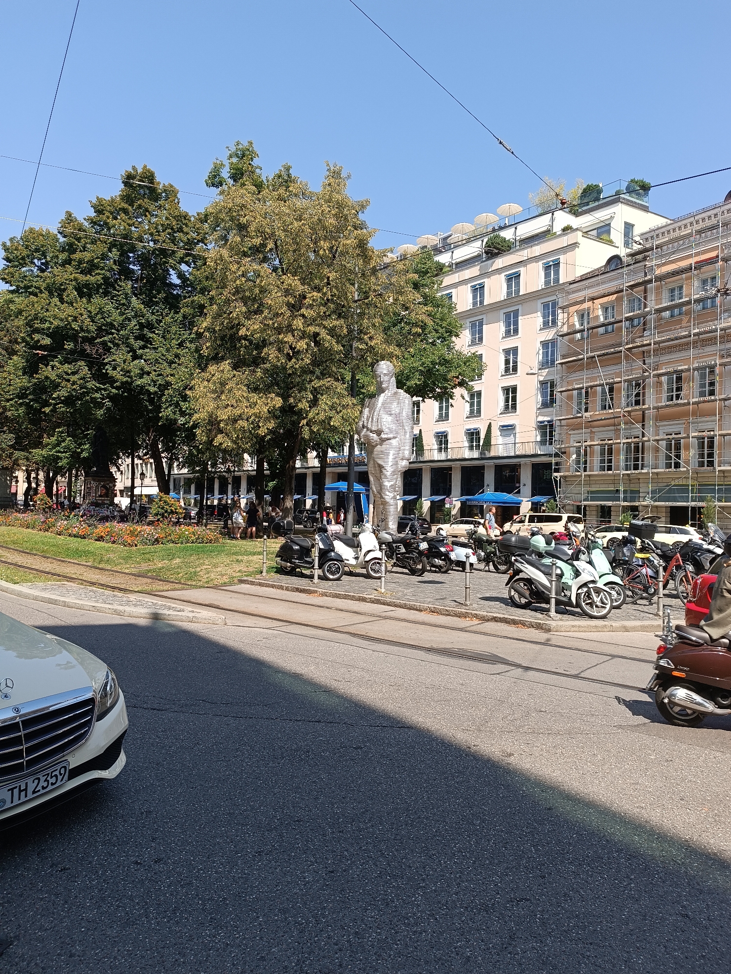 A statue of a man is located in the center of the image. The statue is made of light-colored material and is standing upright. There are several buildings in the background, some of which are under construction. There are also several scooters and a car parked in the foreground. The sky is blue and clear. The scene appears to be a public square in Munich, Germany, likely Marienplatz due to the presence of a statue and buildings characteristic of the area.