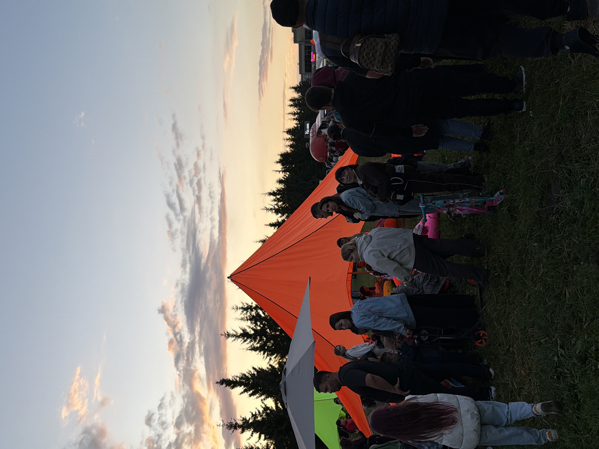 In an outdoor grassy area, a crowd of people gathers in the dimming light of a sunset. A large, bright orange tent dominates the center of the frame, its peak reaching towards the sky. To the left, a large grey umbrella shades part of the gathering, and behind it, a glimpse of a green canopy is visible. Several trees, silhouetted against the colorful sky, form a backdrop. People of various ages are standing, some appear to be conversing, while others are looking around. A child is seen with a scooter, and another is pushing a stroller. In the distance, more trees and a few buildings are faintly visible. The overall atmosphere suggests a community event or festival taking place at dusk.