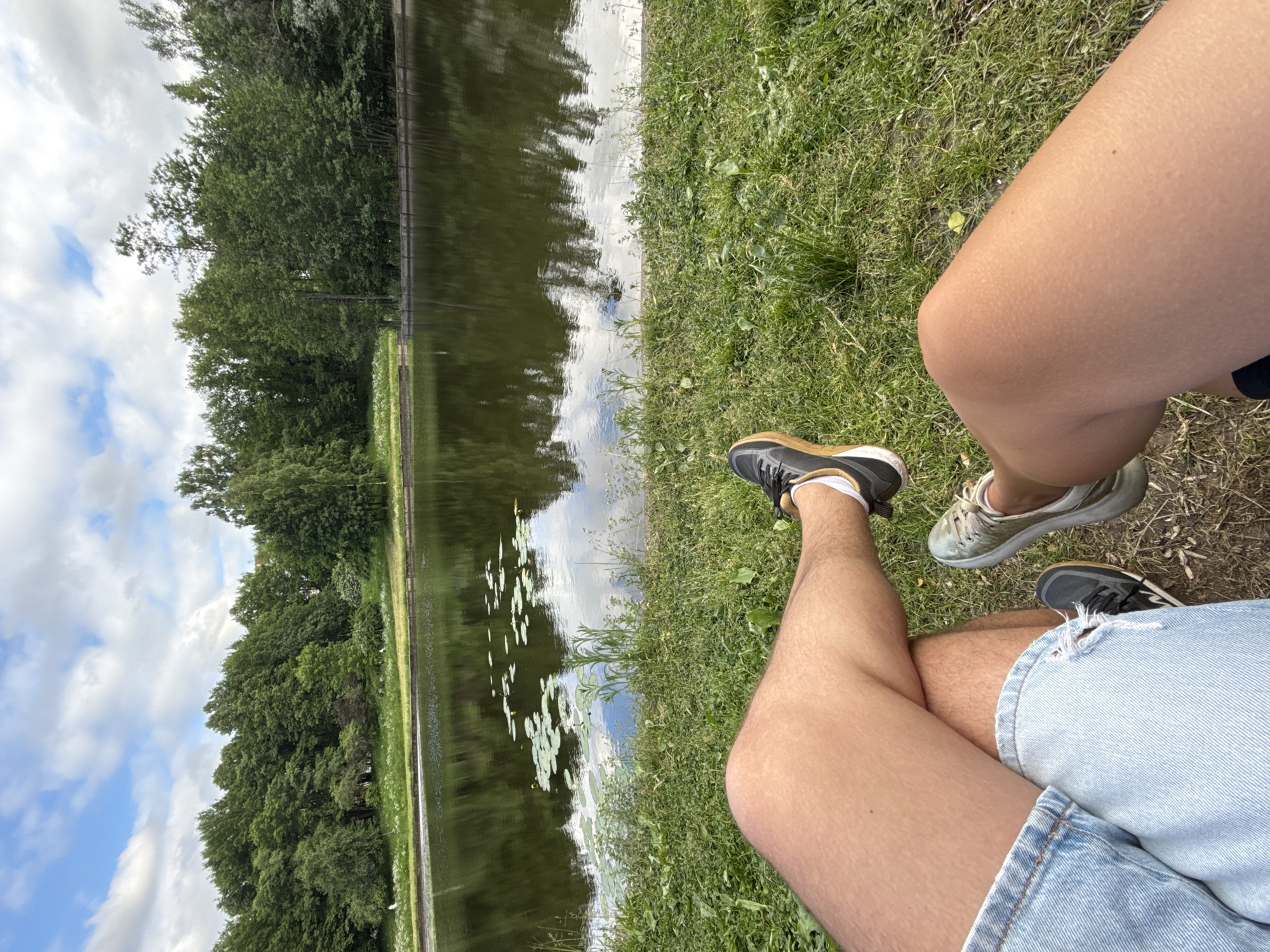 This is a point-of-view shot of two people's legs from the knees down, sitting on a grassy bank overlooking a calm body of water. Both individuals are wearing casual footwear. The person on the left wears light blue denim shorts and black sneakers with white socks. Their legs are crossed, with the right leg on top. The person on the right wears dark shorts and light-colored athletic shoes. Their legs are extended forward and slightly angled. The water in front of them reflects the surrounding greenery and a cloudy sky. Lily pads are visible floating on the surface of the water. The background consists of lush green trees and vegetation lining the far bank of the water. The scene is set outdoors in a park-like environment.