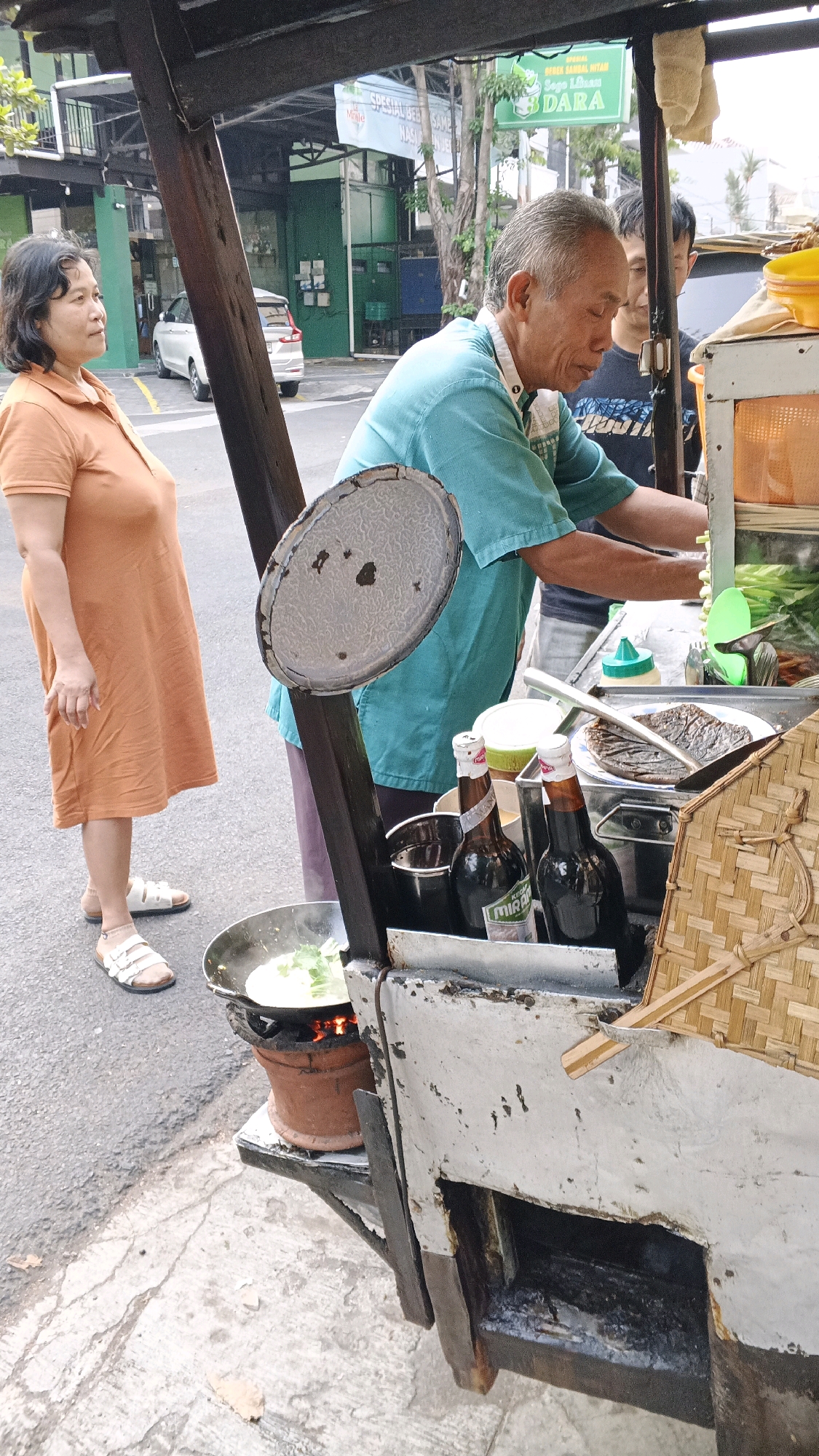 A street food vendor in Semarang, Indonesia is preparing food at a roadside stall. The vendor, an elderly man wearing a teal shirt, is working behind a cart with various cooking implements and ingredients. In front of him, a wok is simmering over a small clay stove. Two bottles of dark liquid, likely soy sauce or a similar condiment, are placed on the cart.
To the left of the vendor, a woman in an orange dress stands watching. She is wearing white sandals. Behind the vendor, another person, a younger man, is visible. The background shows a street scene with buildings, trees, and a parked white car, suggesting an urban environment. A sign in Indonesian is visible above the vendor's stall. The overall scene depicts a typical street food operation in the bustling city of Semarang.