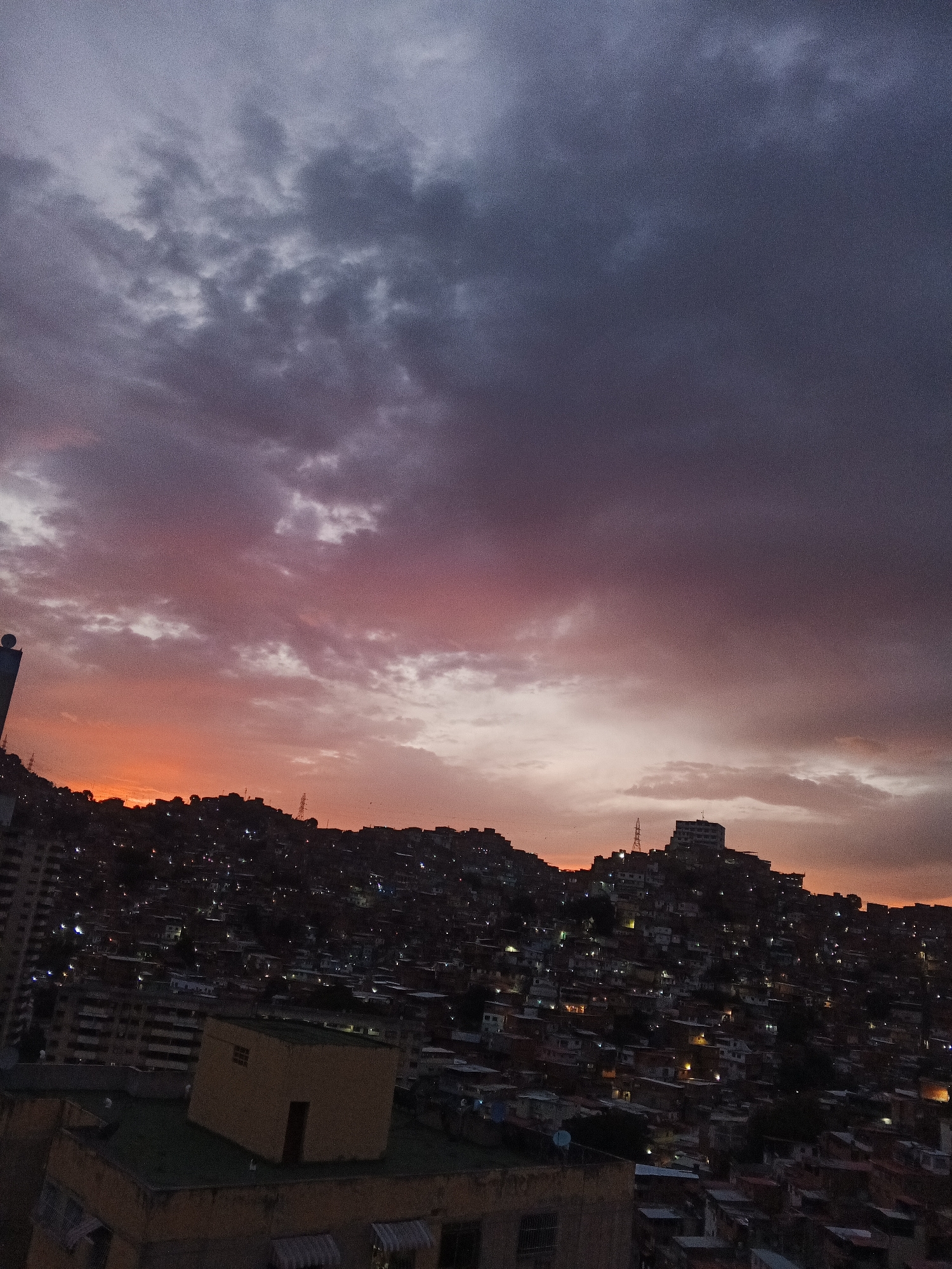 The image depicts a cityscape at twilight in Caracas, Venezuela. The sky transitions from a dark, purplish-gray at the top to a vibrant orange and pink near the horizon. Below, a densely populated hillside is visible, covered in numerous small buildings and houses. Lights from the buildings twinkle, creating a network of illuminated points across the slope. In the foreground, rooftops of nearby buildings are visible, suggesting the photograph was taken from an elevated position. A tall building is visible on the left. The skyline is silhouetted against the colorful sky, with the top of the hill featuring a tower and larger structure that stands out.