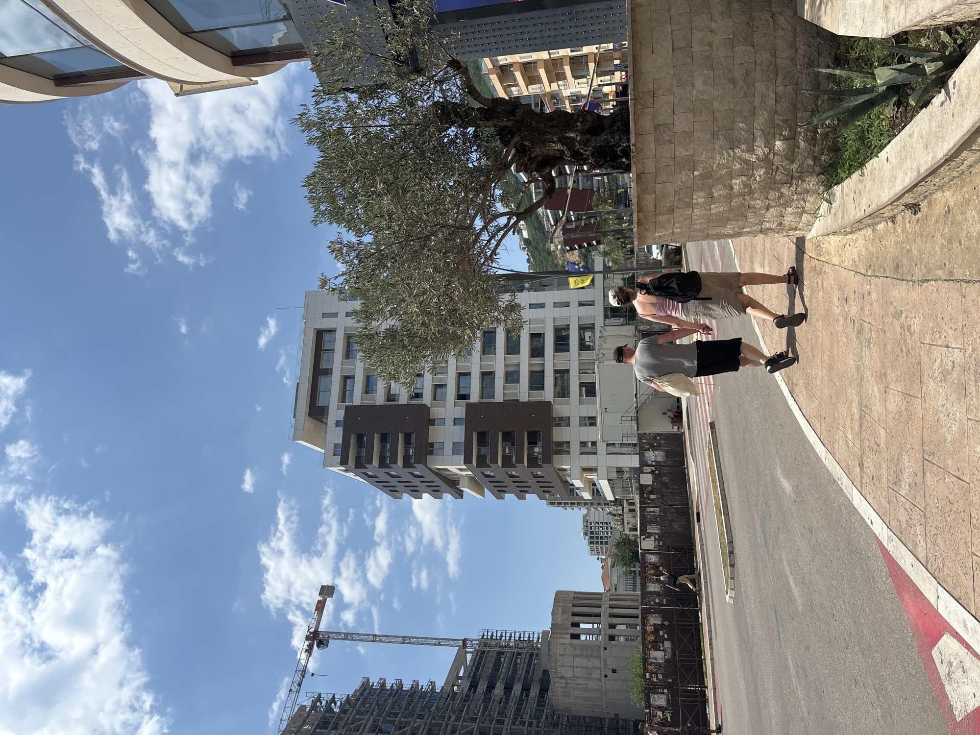The image shows a street scene in Budva, Montenegro, on a sunny day with a partly cloudy blue sky. In the foreground, a couple walks hand-in-hand along a paved sidewalk. The man wears a gray t-shirt and black shorts, while the woman is in a pink tank top and a beige skirt, carrying a black backpack. They are walking away from the viewer, towards a large, mature olive tree with a thick, gnarled trunk, situated on a raised stone planter.
Behind the tree, a multi-story modern building with white and brown facades dominates the mid-ground. To the left of this building, a large construction site is visible, featuring a tall crane and scaffolding on a partially constructed building. Further in the background, more buildings and hills can be seen.
The street in front of the sidewalk appears to be a road with painted lanes, including a red and white marking. Across the road, a fenced-off area with some posters is visible, with a few people scattered around. The overall environment suggests an urban setting undergoing development, likely in a coastal area given the architectural styles and the presence of olive trees.