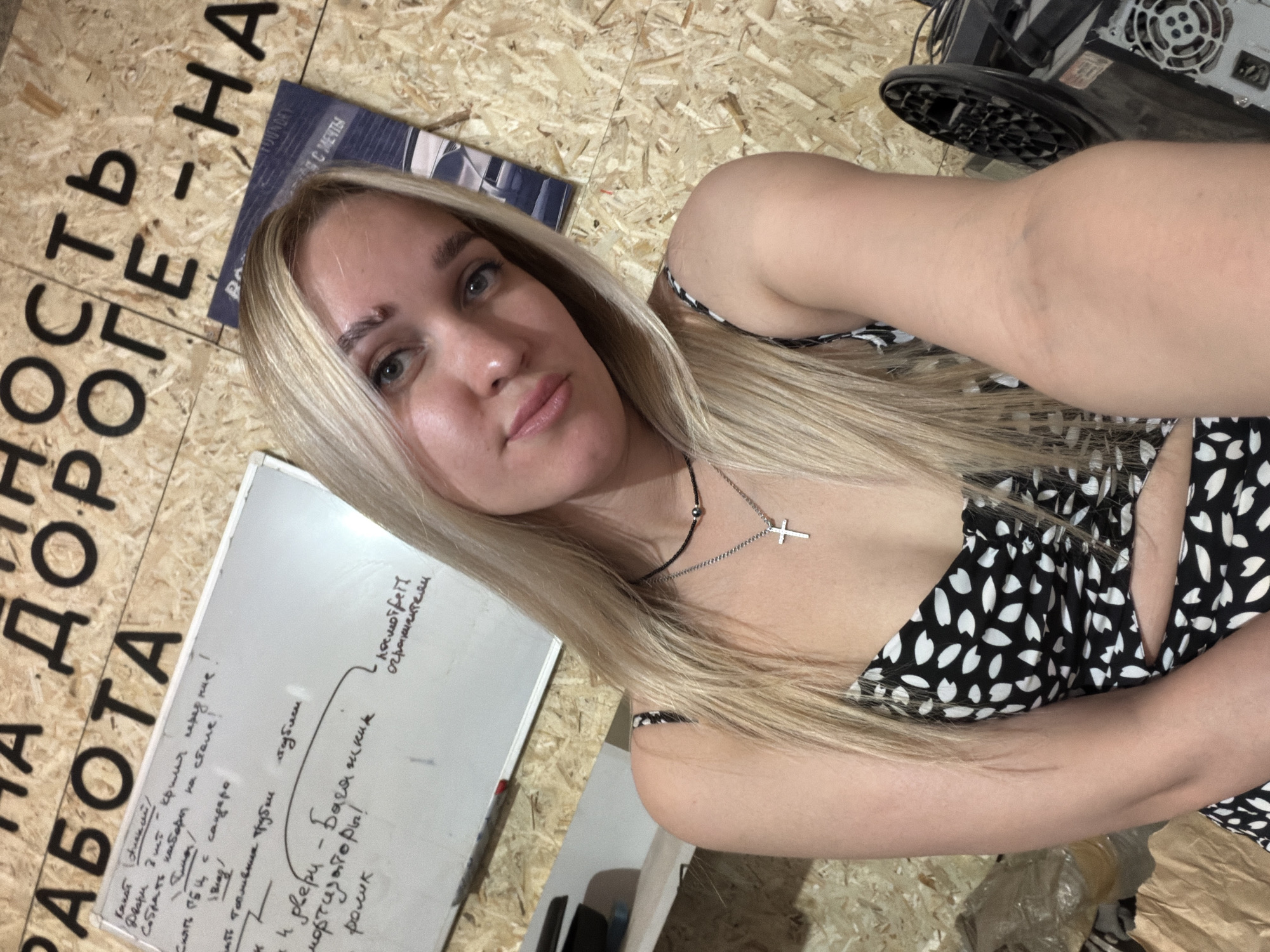 The image depicts a blonde woman with light eyes and a cross necklace lying on a wooden surface with text written in Cyrillic script. There is a white board with handwriting on it nearby, along with what appears to be a magazine or book cover. A piece of machinery is visible in the upper right corner of the frame.