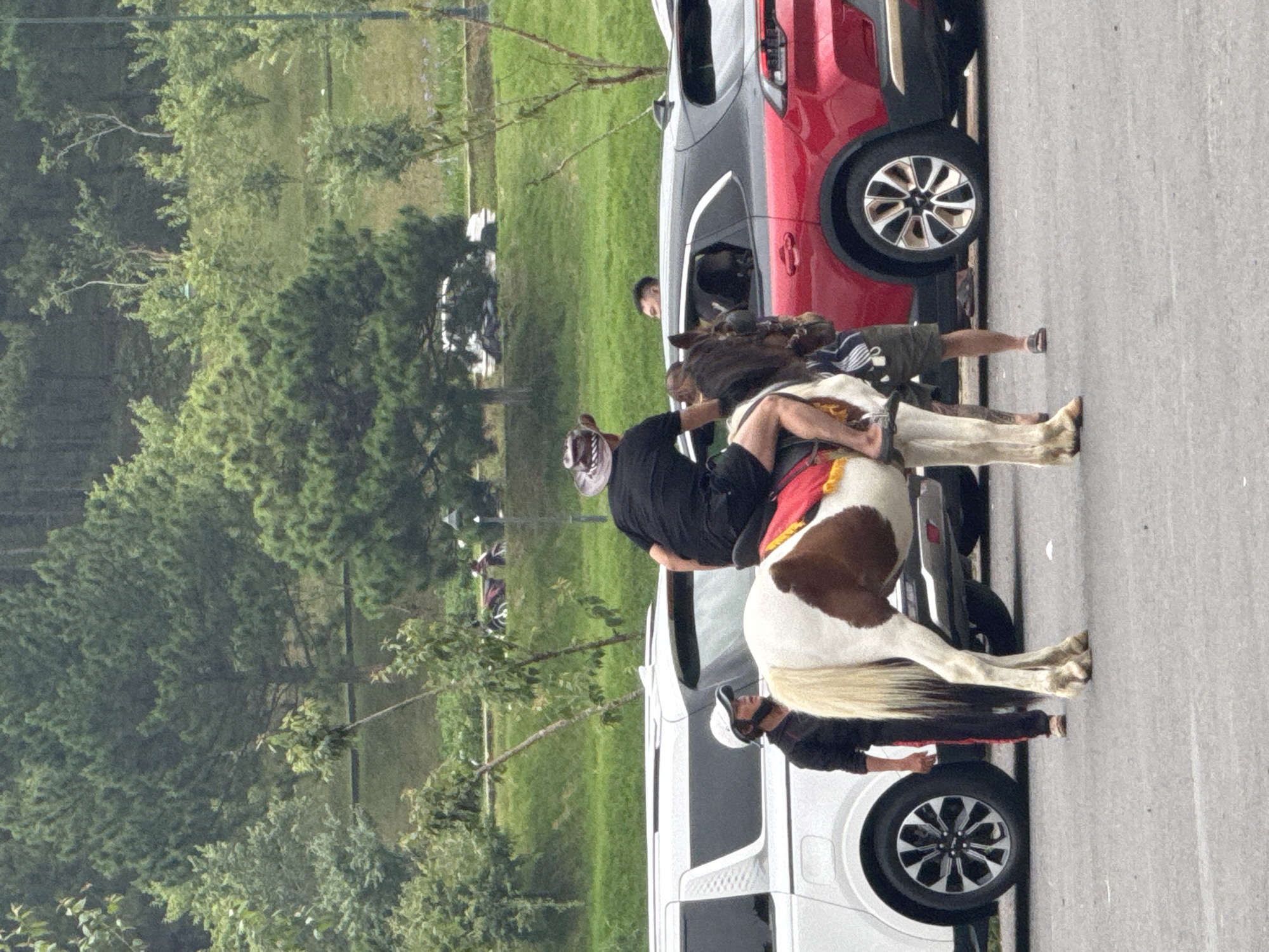 In Phường Xuân Hương, Đà Lạt, Vietnam, a man wearing a black t-shirt and shorts, along with a cowboy hat, is riding a piebald horse. The horse is white with brown patches and has a red saddle blanket. To the left of the horse, a person in a black jacket and white cap is standing. In the background, a white SUV and a red SUV are parked. The scene appears to be on an asphalt surface with a grassy, tree-covered hillside behind the vehicles.