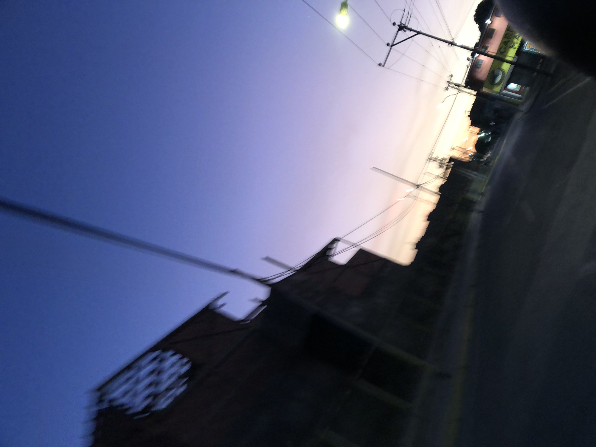 The image depicts an urban scene in Juan Griego, Venezuela, taken at dusk or dawn. The perspective is tilted, showing a street with buildings and utility poles against a gradient sky transitioning from blue to a lighter hue near the horizon. A dark, partially obscured building with an architectural feature resembling a rose window is visible in the foreground. Utility lines stretch across the sky. A street light is illuminated. A building with signage is visible in the distance. The image is blurry, suggesting motion or a fast shutter speed.