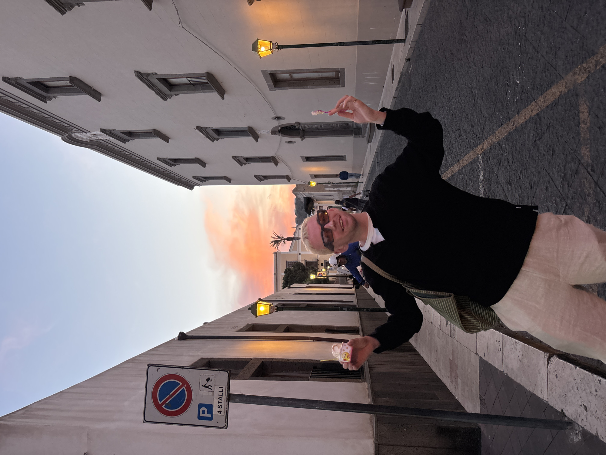 The image, which is rotated 90 degrees clockwise, captures a man standing on a narrow street in Lipari, Italy, during what appears to be sunset or dusk. The man is in the foreground, smiling and looking directly at the camera. He wears dark sunglasses, a black long-sleeved shirt, and light-colored trousers. In his raised right hand, he holds a small, light-colored cup with a spoon, likely containing ice cream or a dessert.
The street is lined by tall, light-colored buildings with multiple dark-framed windows, some featuring decorative ledges and cornices. Several traditional streetlights, illuminated with a warm yellow glow, are visible along the street and against the buildings. In the background, the sky displays vibrant orange and pink hues, characteristic of a sunset or sunrise, with a palm tree silhouetted against the colorful horizon. Other blurred figures of people can be seen further down the street.
The street surface is dark asphalt with faint yellow and white markings. A prominent white road sign with a dark pole stands in the lower left foreground. The sign features a red circle with a diagonal line (indicating no parking or stopping), a blue 'P' for parking, an icon of a person with a cane (disabled parking), and the Italian text "4 STALLI". The overall scene conveys a cheerful and relaxed atmosphere, suggesting an enjoyable evening in a Mediterranean town.