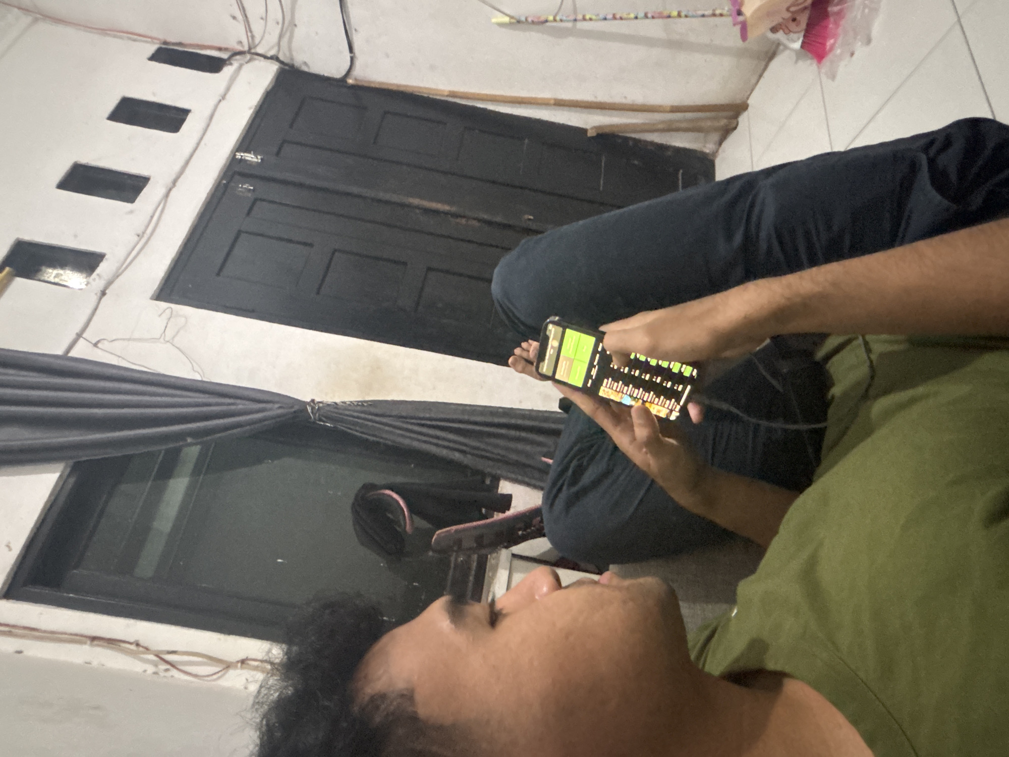This image captures an indoor scene, likely in North Jakarta, Indonesia, during the evening or night. The primary subject is a person, likely male, lying on their back or side, visible from the chest up with dark, curly hair and a relaxed expression. This person is wearing a green t-shirt.
In the mid-ground, another person's (or the same person's) lower body is visible, clad in dark blue pants. An arm extends forward, holding a black smartphone with both hands. The phone screen is illuminated, displaying a grid-like interface with green and white elements, suggesting a game, a music sequencer, or a financial trading application. Earphones appear to be connected to the phone, with a wire visible near the hands.
The background reveals parts of a room. To the left, a dark, heavy curtain or fabric is draped, possibly covering a window. Below this, a dark-framed window is partially visible. Further back and to the right, a dark, two-panel wooden door with a silver latch is seen. The walls are painted white, and above the door, several rectangular recesses or vents are cut into the wall. Various wires, some red and white, run haphazardly along the wall and floor. A wooden stick or rod and a white wire clothes hanger are also visible on the floor near the door. The floor appears to be a light-colored tile or concrete.
The activity central to the image is the interaction with the smartphone. The overall atmosphere is casual and relaxed, with artificial lighting casting soft shadows. No discernible text is visible on the phone screen or elsewhere in the image.
