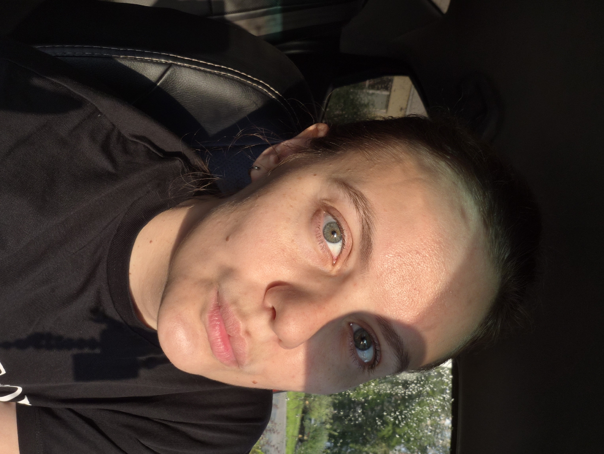 A close-up, head-and-shoulders portrait shows a young woman with light skin and medium-length brown hair pulled back. She is seated in what appears to be a car, with the black leather of a car seat visible on the left side of the frame. The woman is looking directly at the camera with a slight pout. Sunlight falls across her face, casting a shadow over the left side of her forehead and cheek. Her eyes are a greenish-blue. She is wearing a black t-shirt. Out the window, trees and foliage are visible, along with what looks like a building. The image is likely taken in Chaikovsky, Russia, given the context.