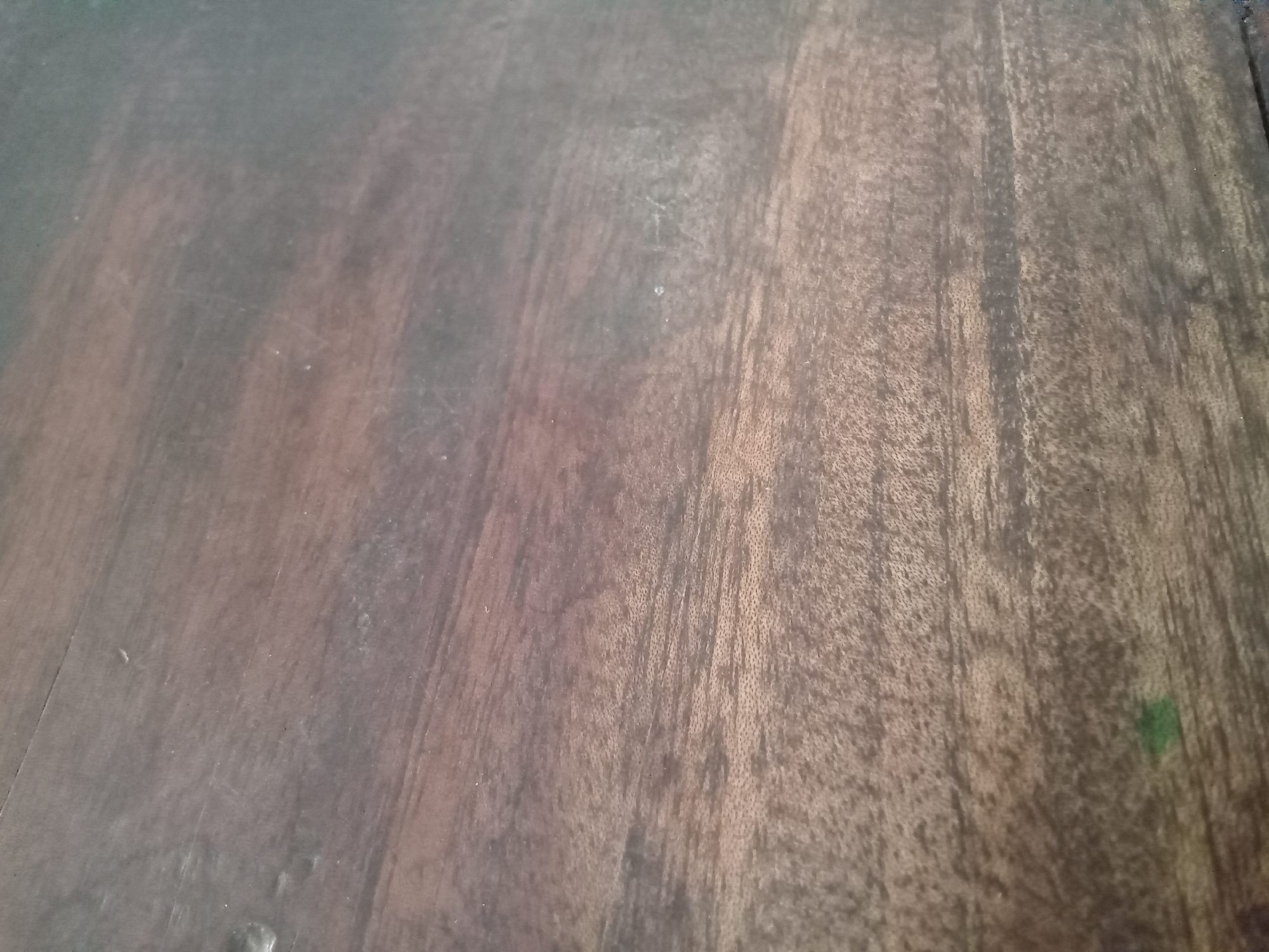The image is a close-up, high-angle shot of a wooden surface. The wood has a dark brown color with visible grain patterns running diagonally from the bottom left to the top right. There are subtle variations in tone and texture, suggesting it might be a table or a floor. Some faint scratches or marks are visible on the surface. In the top left corner, there's a darker, almost black area, and in the bottom right, a small patch of muted green color is present. The lighting is somewhat diffused, creating soft shadows and highlights that emphasize the texture of the wood. The scene does not contain any people, actions, or specific objects that would allow for localization within Simo, Indonesia.