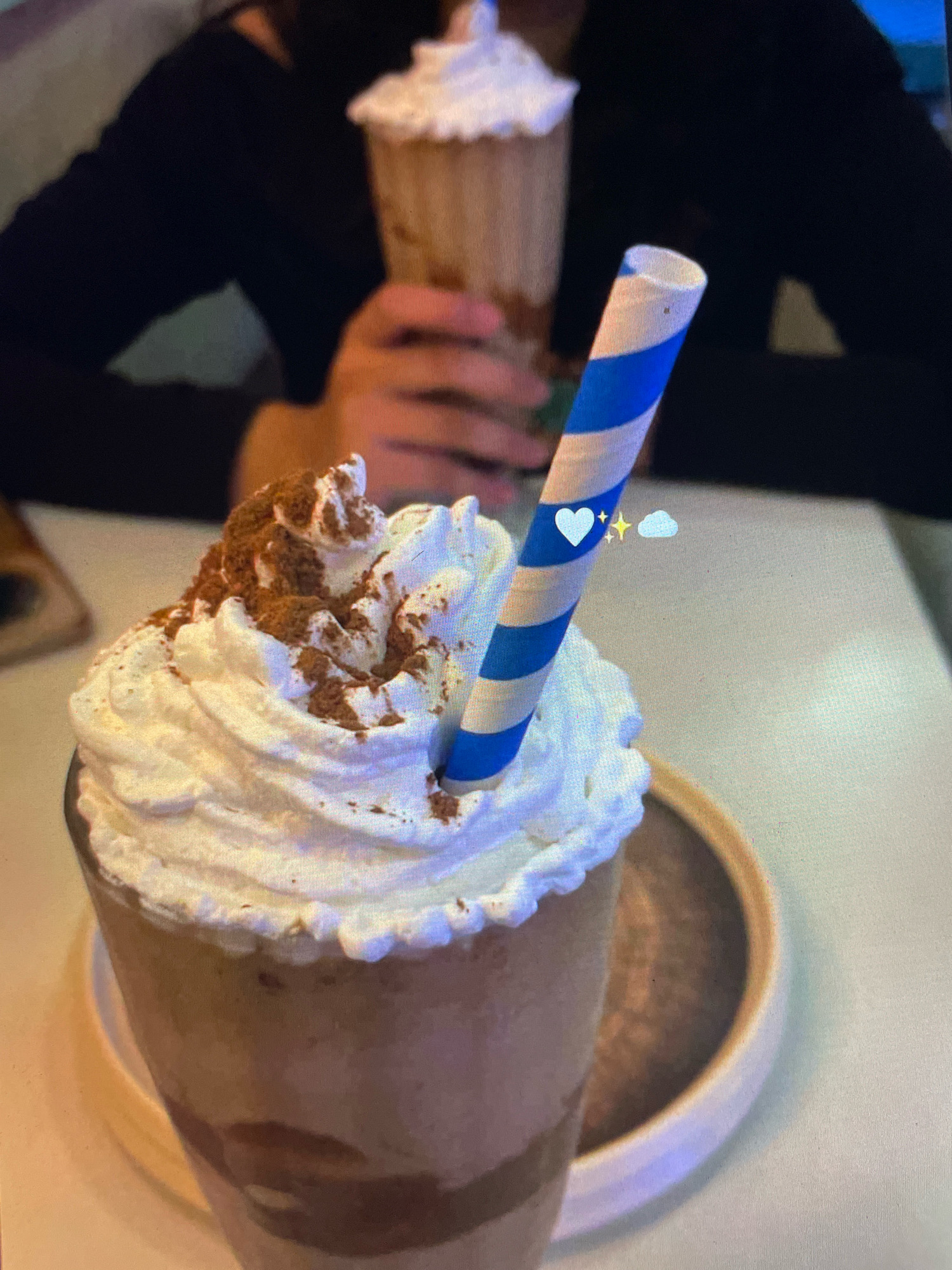 The image depicts a close-up view of a beverage, likely a milkshake or frappe, topped with whipped cream and sprinkled with cinnamon. A blue and white striped straw is inserted into the drink. In the background, a person is visible wearing a dark top, and another blurred beverage is in the distance. The setting appears to be indoors, possibly a cafe. The image does not contain any information that can place it in Gankar, India or any other specific city.