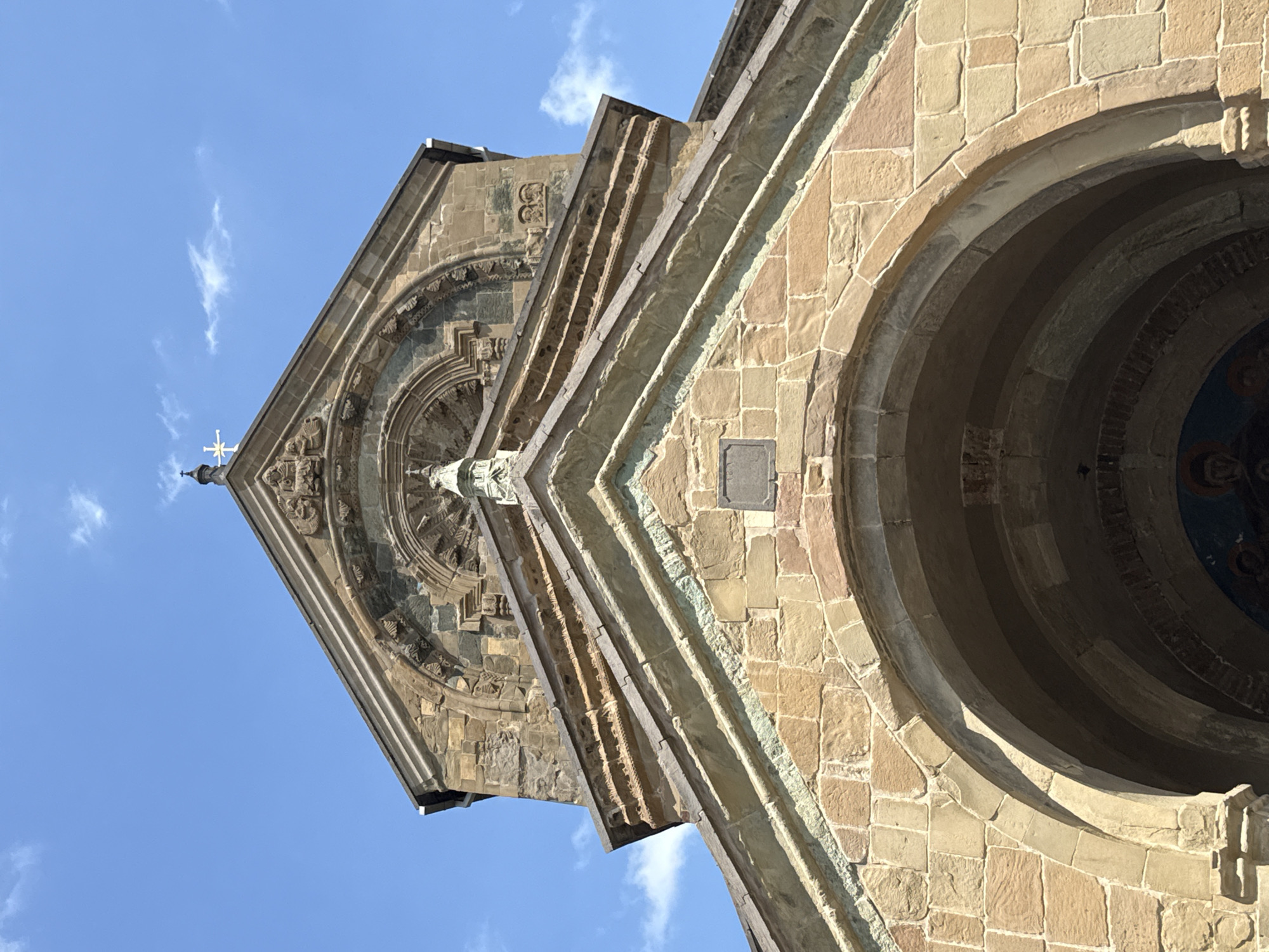 The image shows a low-angle, upward view of the facade of a stone church, likely Jvari Monastery, located on a hill overlooking Mtskheta, Georgia. The architecture is characterized by weathered, light brown and tan stone blocks, with decorative carvings and a prominent arched entryway at the bottom. A cross tops the triangular gable of the roof. The sky above is a clear blue with a few wispy white clouds. The scene is static, with no people or active events depicted.