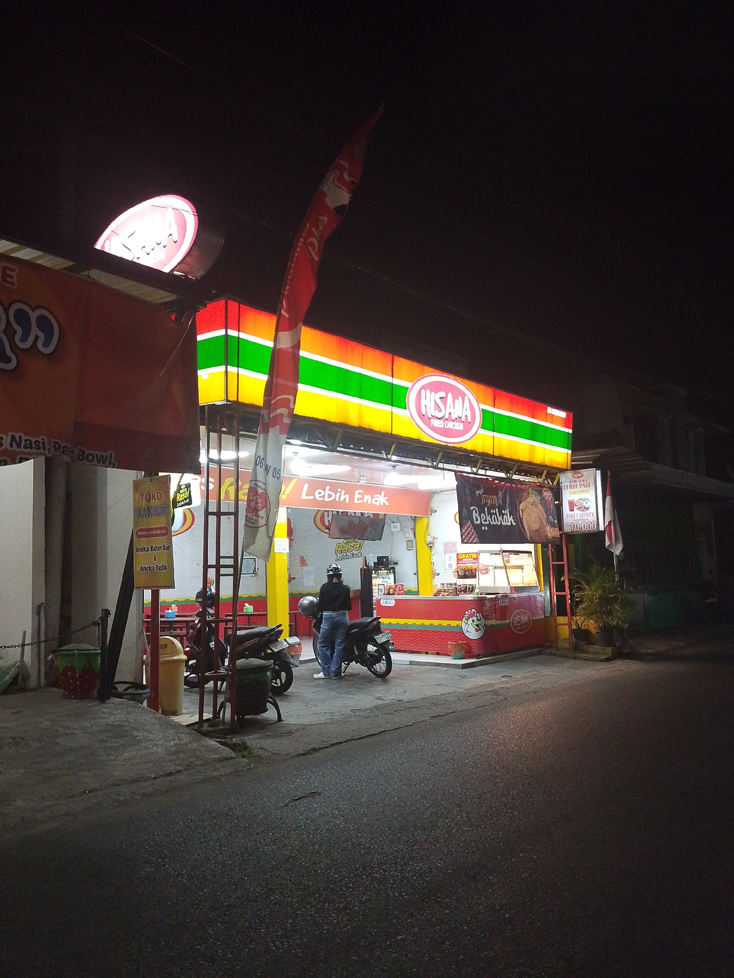 The image captures a nighttime scene outside a Hisana Fried Chicken restaurant in Kediri City, Indonesia. The restaurant features a brightly lit facade with a prominent "HISANA FRIED CHICKEN" sign and colorful striped lighting. A banner inside the restaurant reads "Lebih Enak" (More Delicious). Several motorcycles are parked in front of the establishment, and a person in blue jeans and a dark jacket stands near one of them, appearing to be a customer or employee. Trash cans and a sign for "TOKO NAKURRY" (Nakurry Store) are visible on the left side of the image. The street in front of the restaurant is wet, suggesting recent rain. The overall atmosphere is that of a typical urban commercial area at night.
