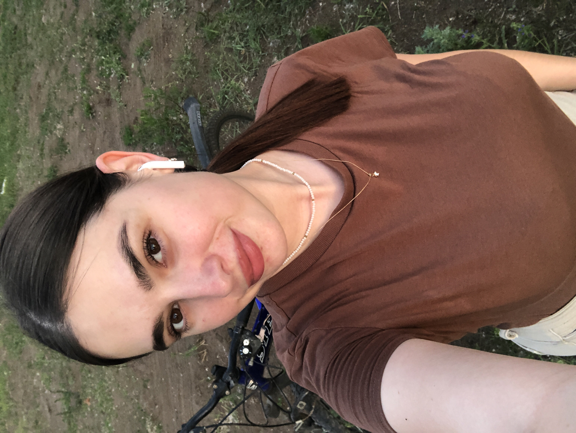 A young woman with dark hair, brown eyes, and light skin takes a selfie outdoors. She is wearing a brown t-shirt, a white beaded necklace with a small gold pendant, and white wireless earbuds. A bicycle is visible behind her, and the background consists of dirt and grass, suggesting a rural or park setting. The image does not provide any specific information to determine the location in the city of Chaikovsky, Russia.