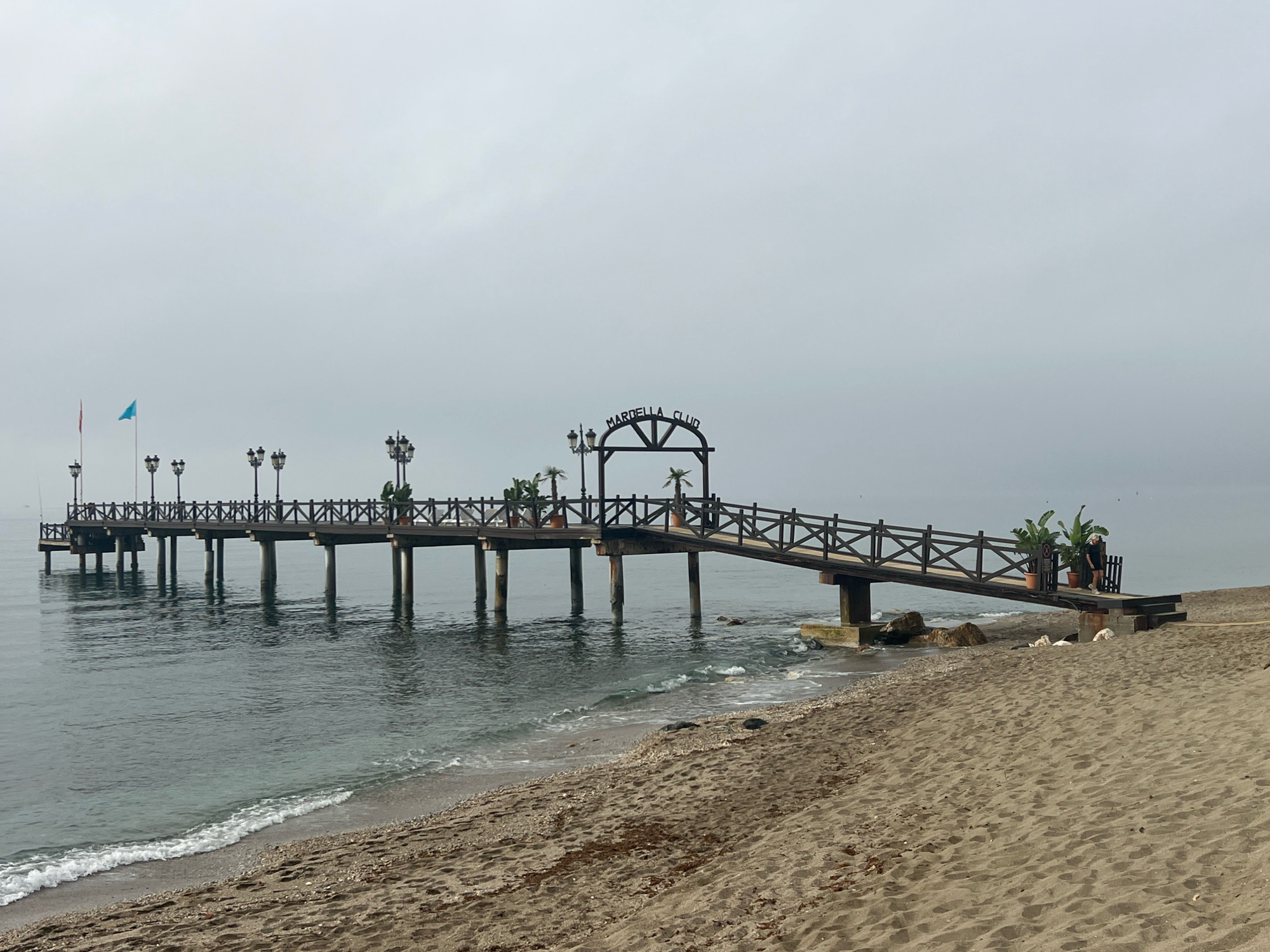 The image shows the pier of the Marbella Club in Marbella, Spain, on a cloudy day. The wooden pier extends out into the calm, gray water of the sea. The pier is lined with streetlights and potted plants, and at the entrance to the pier, there is an archway with "MARBELLA CLUB" written on it. A sandy beach with some rocks and seaweed is in the foreground, and a few waves are breaking on the shore. A woman is standing on the pier, looking out at the sea. The overall atmosphere is serene and tranquil.