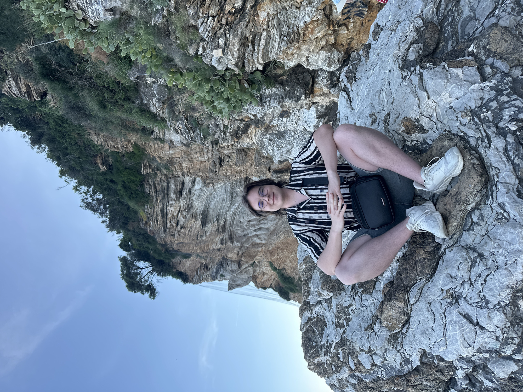 A person is sitting cross-legged on a rocky outcropping. They are wearing glasses, a black and white striped shirt, and gray shorts. A black bag is on their lap. The person is wearing white sneakers. The rocky area overlooks a cliffside and the sea. The image was taken in Budva, Montenegro.