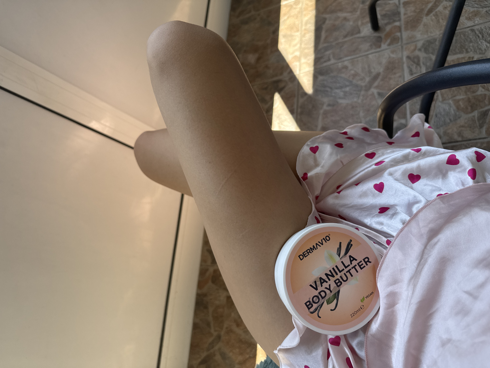 The image displays a close-up, low-angle view of a person's crossed legs, situated in what appears to be a residential interior or balcony area, potentially in Plovdiv, Bulgaria. The bare legs exhibit a light skin tone. The person is wearing light-colored fabric, likely shorts or pajamas, embellished with small red heart patterns. Centrally placed on the fabric between the legs is a round tub of "DERMAV10 VANILLA BODY BUTTER," featuring a white lid and a peach-hued label that includes text indicating "VANILLA BODY BUTTER," "220ml e," and "VEGAN," alongside illustrations of vanilla beans and a flower. To the lower right, a curved segment of a black, matte-finished metal chair frame is visible. The background consists of a light-toned ceiling or upper wall in the top portion, traversed by a slender dark line. The lower background reveals a tiled floor composed of irregular, earthy-toned tiles (shades of brown, grey, and terracotta). Prominent, sharply defined shadows are cast across the tiled surface by strong natural light, indicating direct sunlight entering the space.