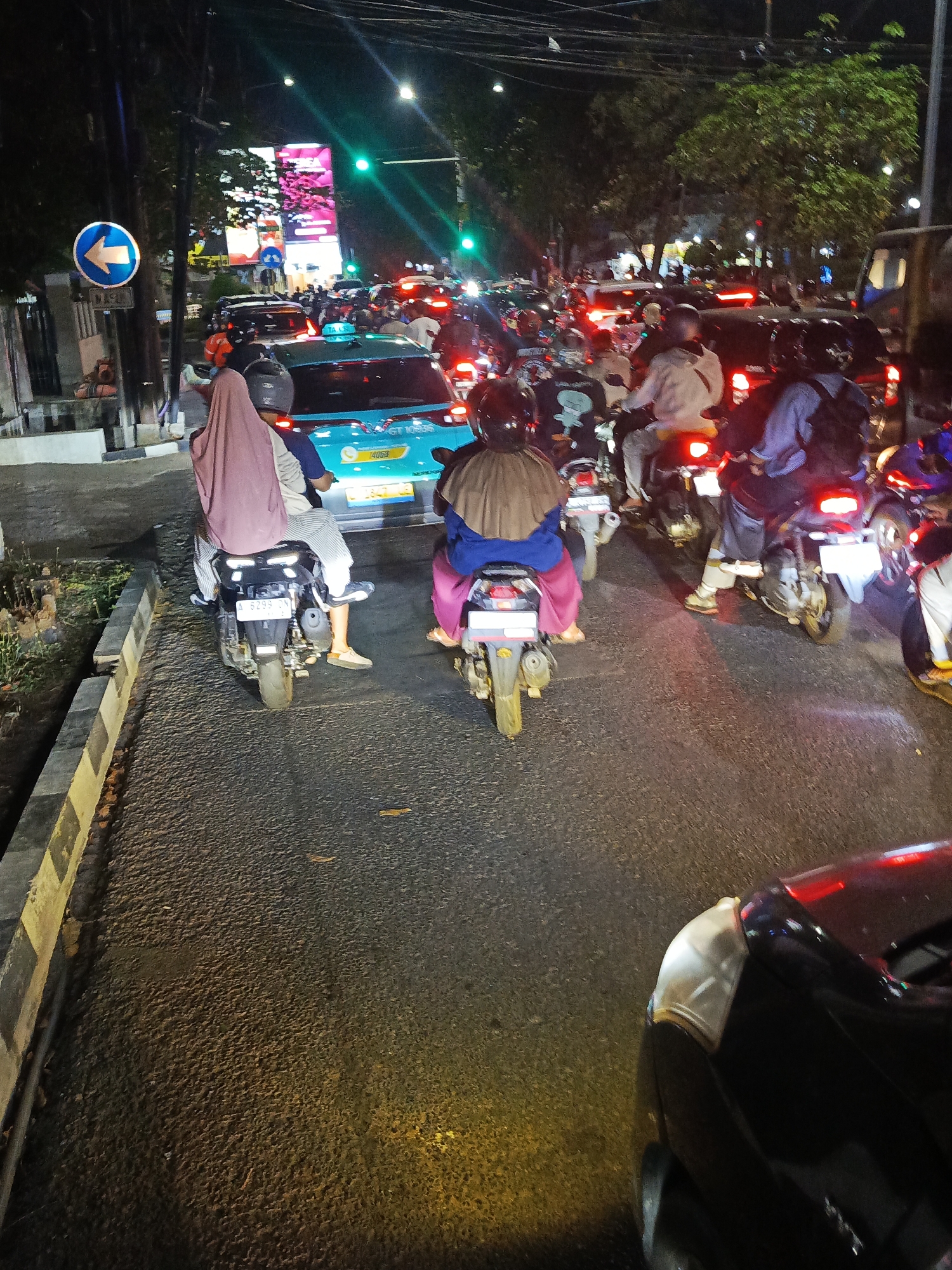 The image depicts a busy street scene in Surabaya, Indonesia, at night. A traffic jam is evident, with numerous motorcycles and a few cars filling the road. The lighting suggests it is evening or night, with streetlights and illuminated signs contributing to the visibility.
In the foreground, the rear of a black car is visible, its brake lights illuminated. To the left, a blue taxi with the number "GT 10595" on its license plate is among the vehicles. Several motorcycles are in motion, with riders wearing helmets and varying attire.
One motorcycle in the middle foreground carries a driver and a passenger, the passenger wearing a pink headscarf and a blue shirt, with striped pants. Another motorcycle, directly behind the taxi, has a rider wearing a brown hijab and a pink outfit. The license plate of this motorcycle reads "A 6299".
The street is paved and appears to be a main thoroughfare, with trees and buildings lining the sides. A blue directional sign with a white arrow pointing left is visible on a pole to the left of the road. Large illuminated billboards are present in the background, suggesting a commercial or entertainment district. The overall atmosphere is one of urban congestion and nighttime activity.
