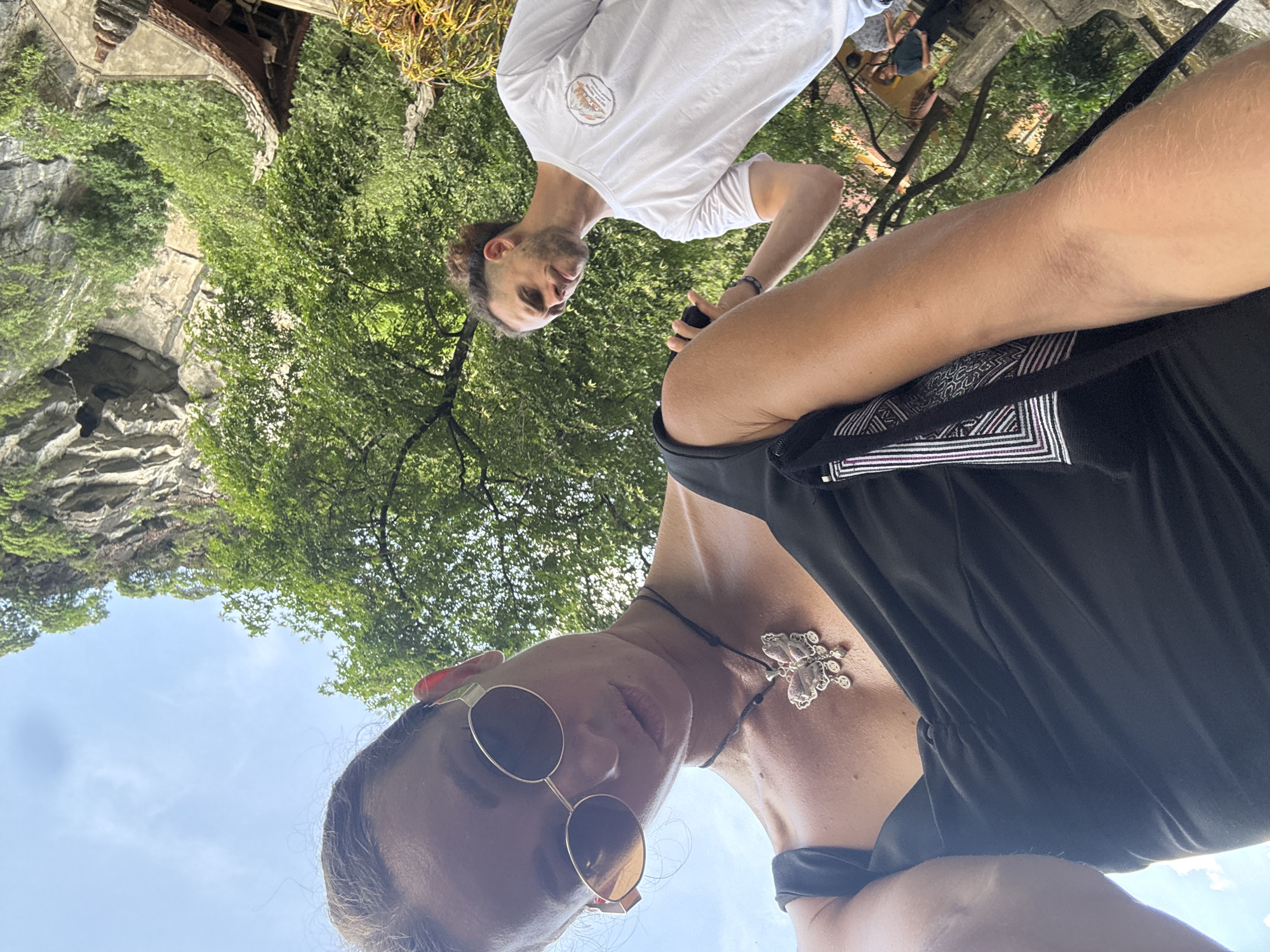 In an outdoor, low-angle selfie, a woman with brown hair pulled back and wearing round, gold-rimmed sunglasses is in the foreground. She is wearing a black, sleeveless dress and a necklace with a butterfly pendant. Behind her, a man with curly brown hair pulled into a bun and a beard is visible, wearing a white t-shirt. He is looking down and to his left, with his right hand raised. In the background, lush green trees and a limestone karst cliff are prominent, suggesting a natural, mountainous environment. A portion of a building with a curved roof, possibly a temple or pagodashaped structure, is visible on the right. The presence of karst formations, lush vegetation, and the architectural style of the building are consistent with the landscape found in Nam Hoa Lư Ward, Ninh Bình, Vietnam, an area known for its scenic karst mountains and ancient temples. Other indistinct figures are also present in the background.