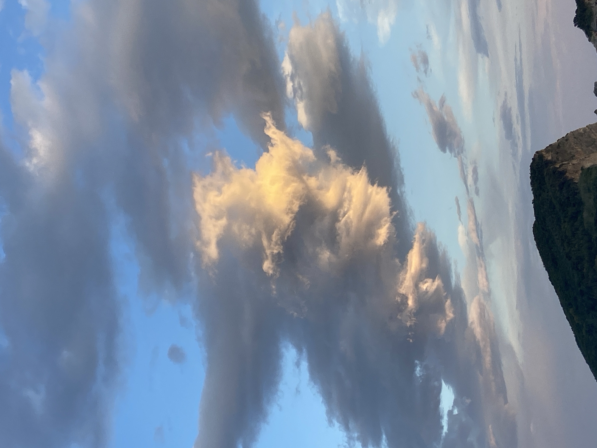The image shows a sky filled with clouds. Some clouds are dark gray, while others are illuminated with a golden hue. The sky itself is a light blue color. Part of a dark green, rocky cliff is visible in the upper right corner. The location and city are unknown.