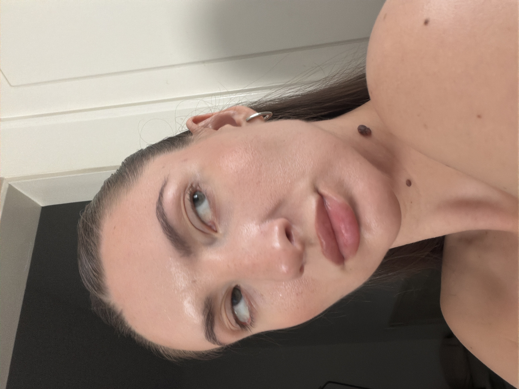 A close-up, high-angle shot features a light-skinned woman with dark hair pulled back, looking to her left. Her face is partially illuminated, highlighting her features and some skin blemishes. She is wearing a hoop earring in her visible ear. The background consists of a dark, out-of-focus area on the left and a light-colored, vertically paneled surface on the right. The image does not depict the city of Sharm El Sheikh, Egypt.