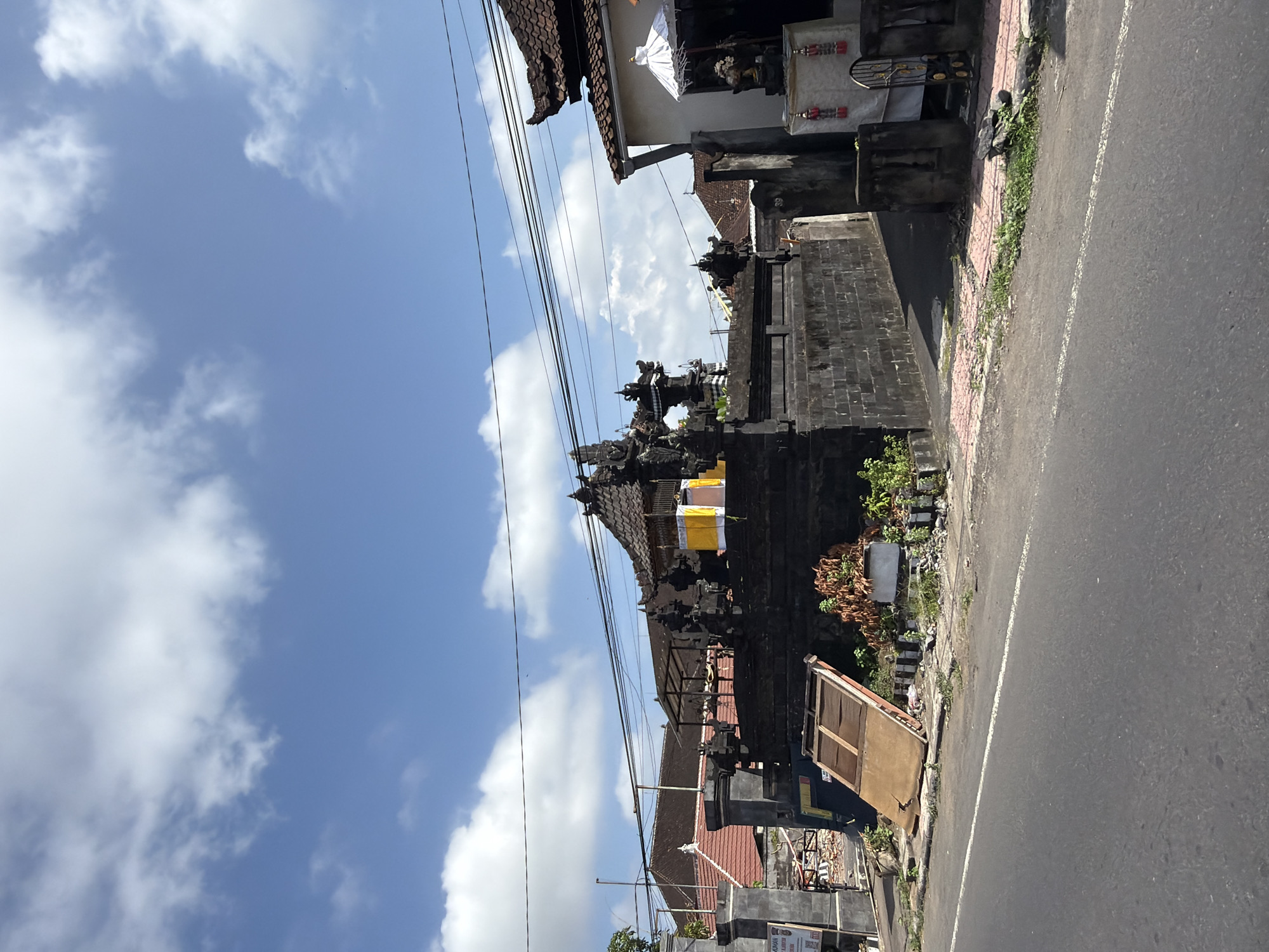 The image shows a street scene in Blahbatuh, Indonesia, on a clear day with scattered clouds. A dark stone temple or shrine complex with elaborate carvings and tiled roofs is the central focus. The complex is bordered by a low stone wall. To the right of the temple, a white umbrella and some religious decorations are visible in front of a building. A street runs in the foreground, with a sidewalk alongside it. On the sidewalk, there is a discarded wooden board and some plants. Overhead, power lines are strung across the sky. The overall impression is of a tranquil, possibly religious, area within a town or village.