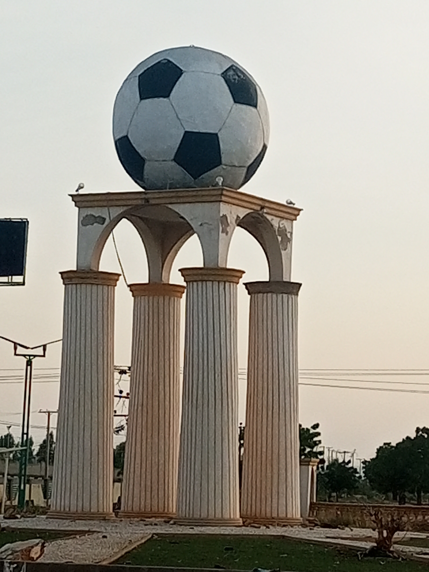 The image shows a large, artificial soccer ball statue atop a structure with four fluted columns and an arched entablature. The statue is white with dark blue pentagons. Two small birds are perched on the top of the entablature. The structure appears to be made of concrete and is located outdoors. In the background, there are trees, utility poles, and power lines, suggesting an urban or suburban environment. The sky is overcast, indicating a possibly hazy or cloudy day. The general setting suggests it's a public landmark or monument. This monument is located in the city of Gombe, Nigeria.