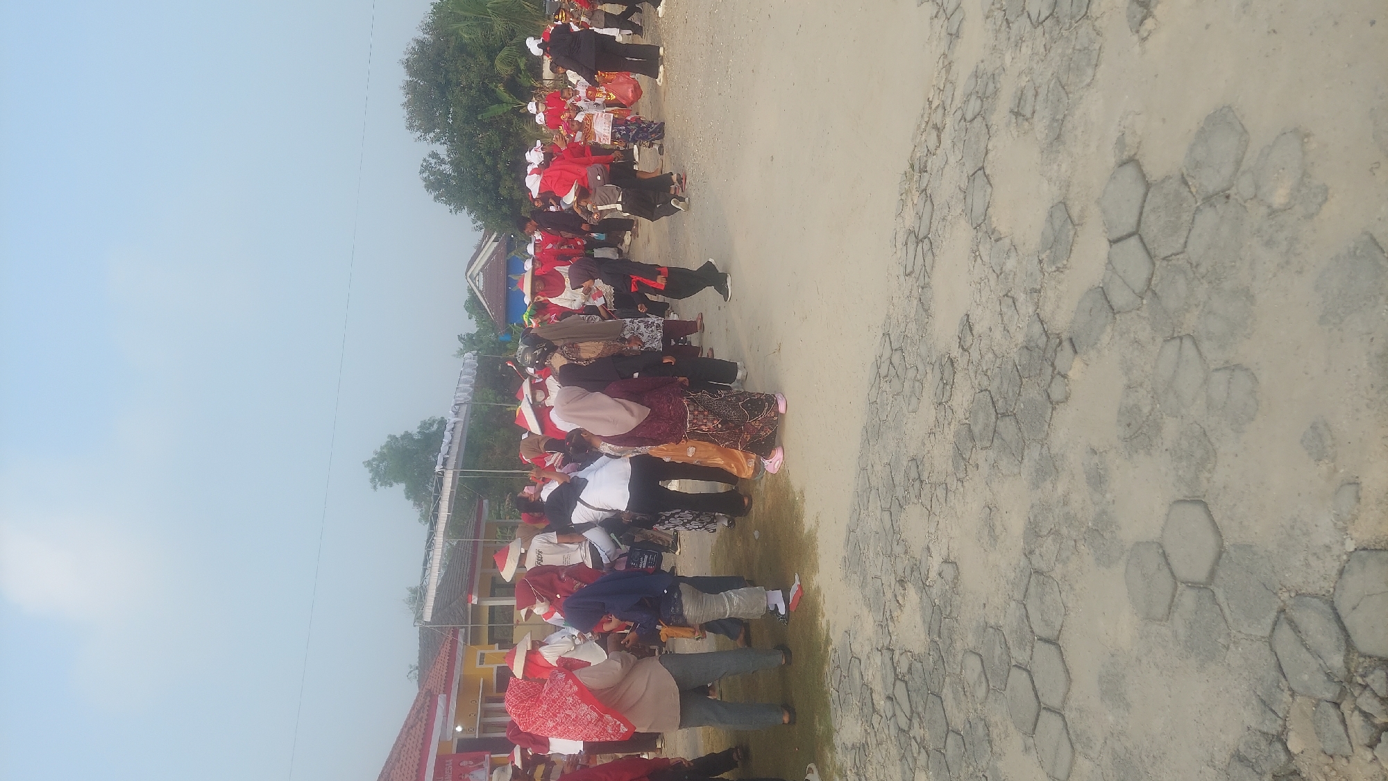 The image captures an outdoor gathering of people, many wearing red and white attire, including hats, suggesting a patriotic event. The setting appears to be a public space with buildings in the background and a ground covered with hexagonal paving stones and dirt. A group of individuals are seen standing and interacting in a relatively open area. The sky is overcast, indicating daytime. The presence of flags and banners suggests a celebratory or ceremonial occasion. The location is identified as Reno Basuki, Indonesia.