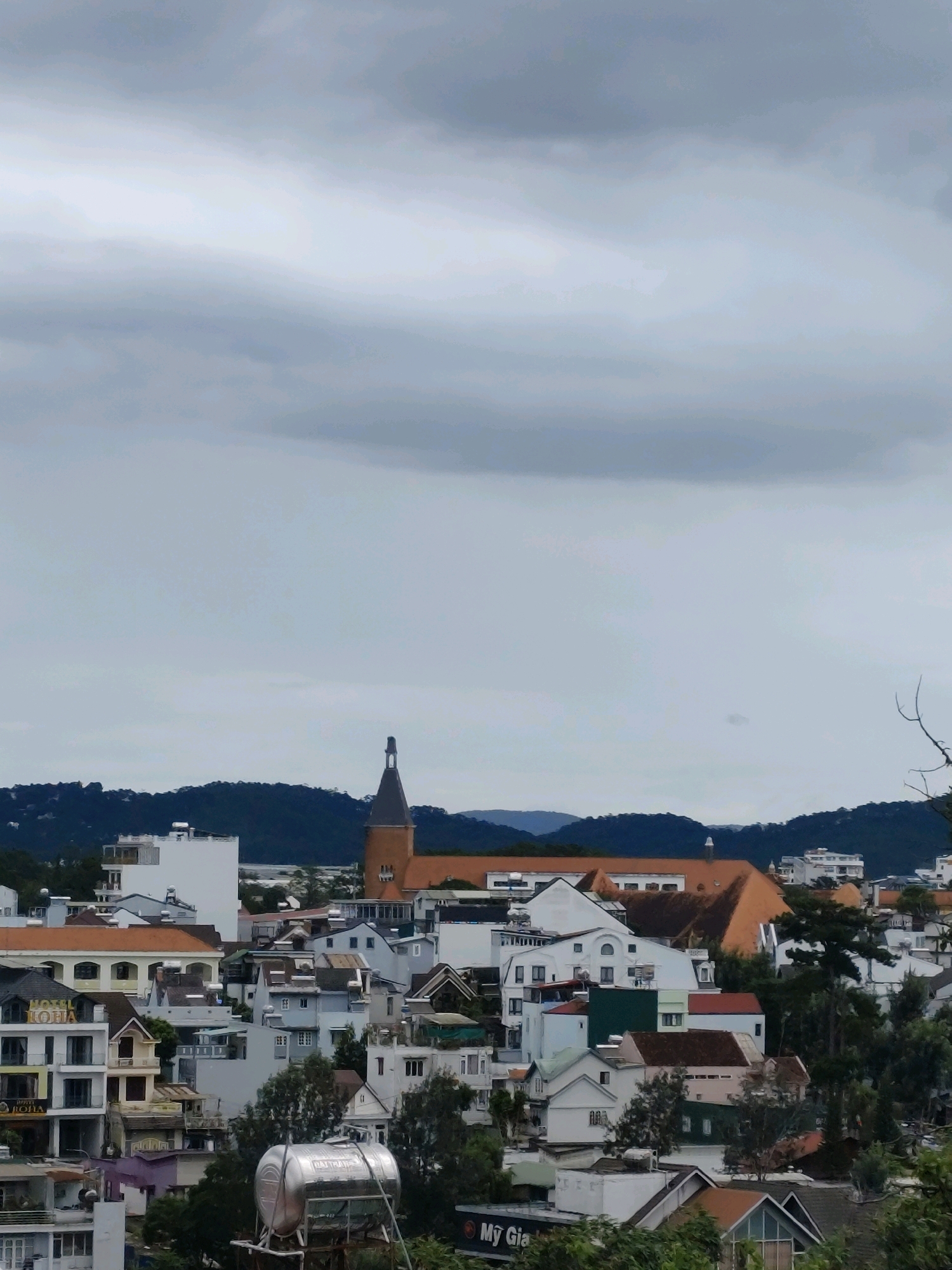 This is a high-angle, medium shot of a city skyline in Da Lat, Vietnam, under a cloudy sky. The city is densely packed with buildings of varying heights and architectural styles, many of which are white or have light-colored facades. In the mid-ground, a prominent brown church steeple with a dark roof stands tall above the surrounding structures. The buildings in the foreground include a white multi-story hotel with signage that reads "IOHA IOHA," a silver water tank on a stand, and various residential buildings with tiled roofs. Lush green trees are interspersed throughout the urban landscape. In the background, rolling, dark green hills form a scenic backdrop. The overall impression is of a vibrant, European-influenced city nestled amongst natural beauty.