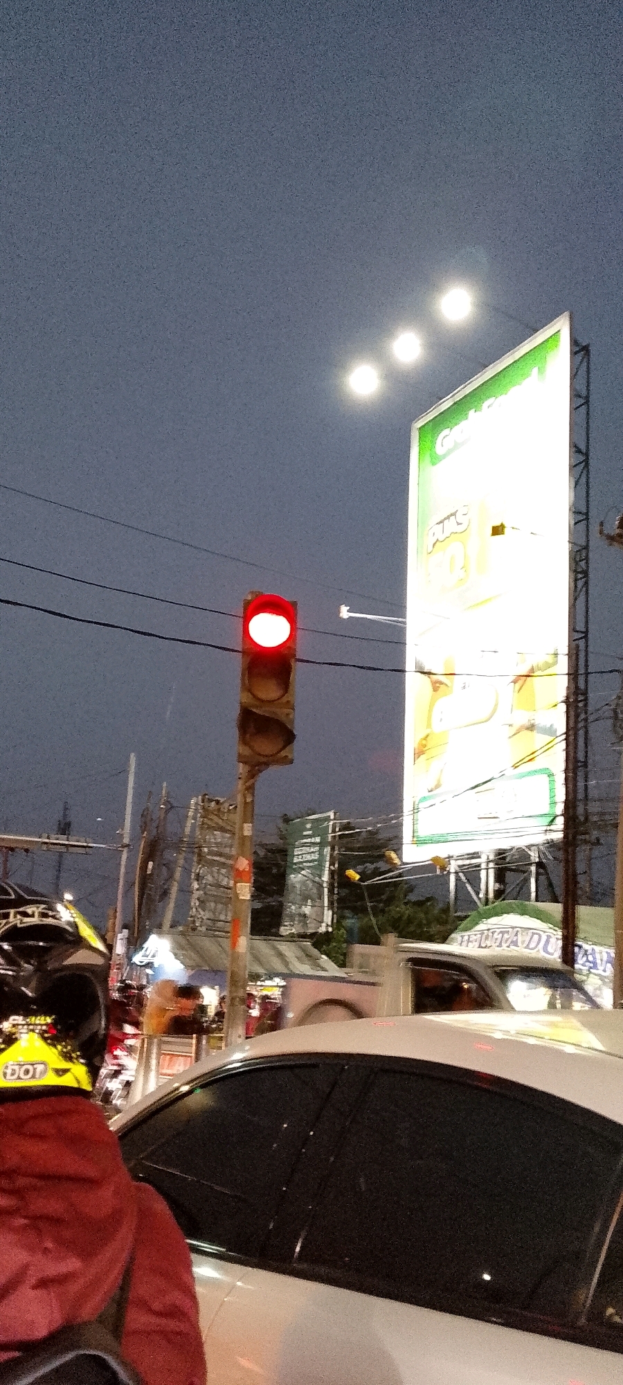 The image captures a street scene in Depok, Indonesia, at dusk. A red traffic light is illuminated, indicating a stop. In the foreground, a person wearing a red jacket and a yellow helmet with a "DOT" sticker is visible. Next to them is the side of a white car. To the right, a large billboard with green and yellow branding, featuring text and images, is illuminated by overhead lights. Several streetlights are also visible in the sky. The background shows a network of power lines and more distant buildings and structures, suggesting a bustling urban environment.