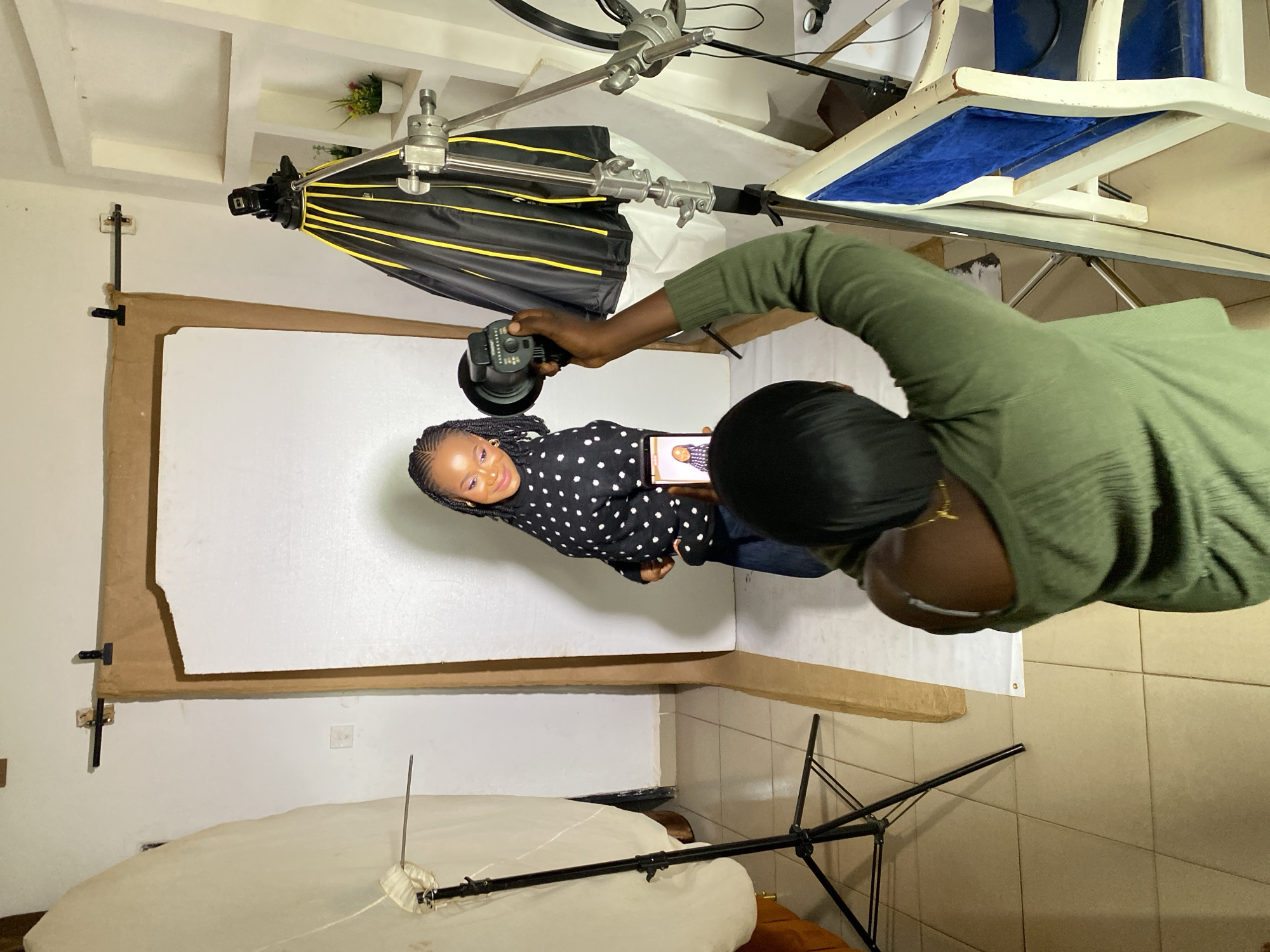 In a studio setting, possibly located in Jos, Nigeria, a photographer, positioned with their back to the camera, is actively taking a photograph of a young woman. The photographer, wearing a green long-sleeved shirt, holds a smartphone in their right hand, aiming it at the subject. The subject, a young woman with braided hair and dark clothing adorned with white polka dots, stands facing the camera with a slight smile. Behind her, a plain white backdrop is used. The studio is equipped with photographic lighting, including a softbox on the left and a flash umbrella on the right, supported by stands. A wooden backdrop frame with brown paper lining the sides is visible. The floor is tiled.